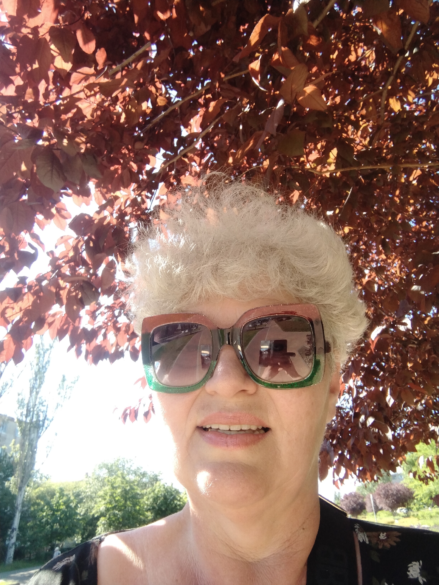 A woman with short white hair and large, colorful sunglasses is standing under a tree with reddish-brown leaves. The sunglasses reflect the scene around her. She is wearing a floral-patterned top. Behind her are trees, a building, and what appears to be a park or green space. The location is Horishni Plavni, Ukraine. The sun appears to be shining brightly, casting shadows on her face and the surrounding environment.