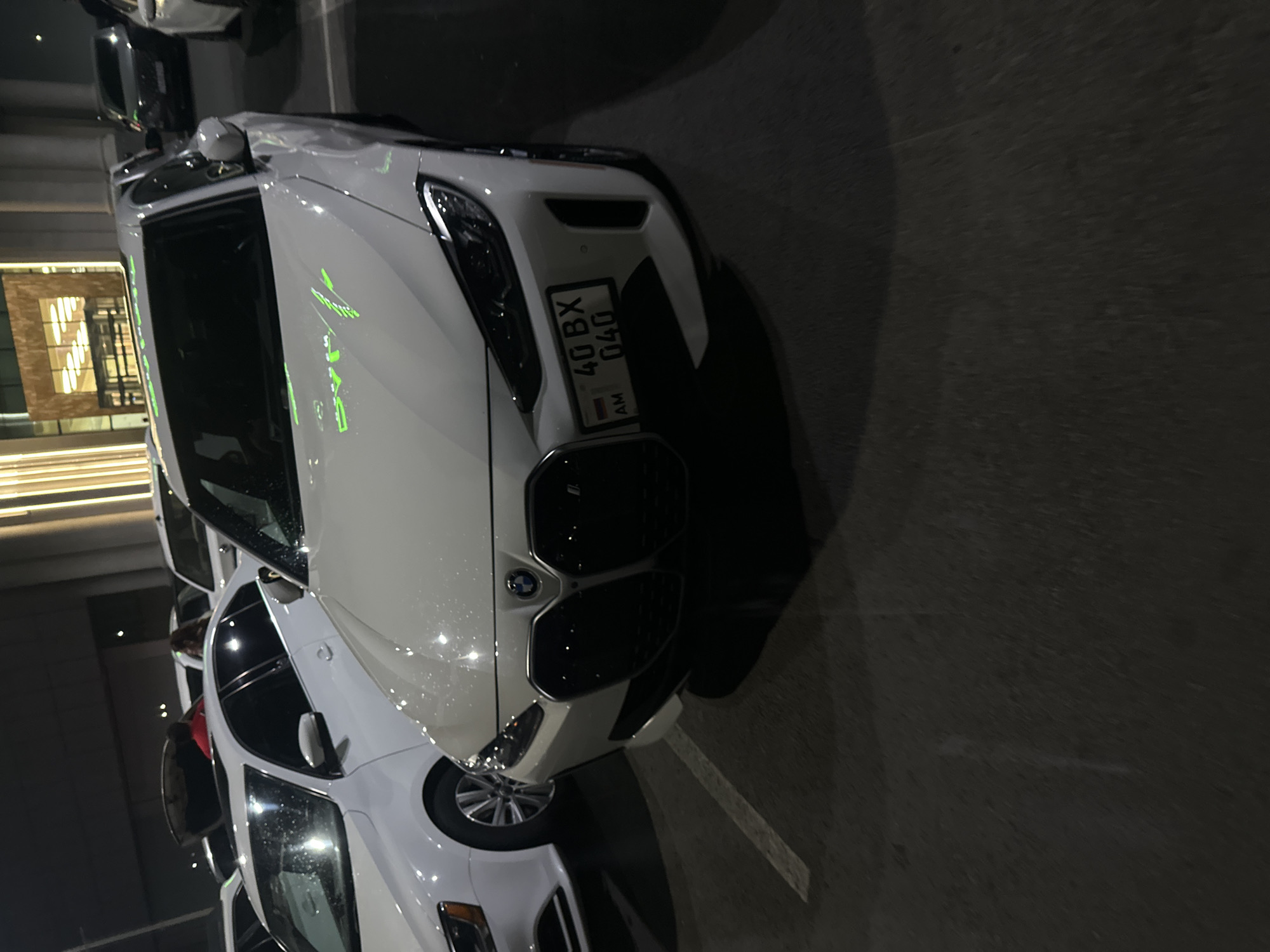 The image shows a white BMW car parked in an unknown city location. The car has a license plate that reads "AM 40 BX 040." The car is situated in a parking area, with other vehicles visible in the background. The environment is an urban setting, possibly at night, with buildings visible in the background. The scene is lit by artificial light.
