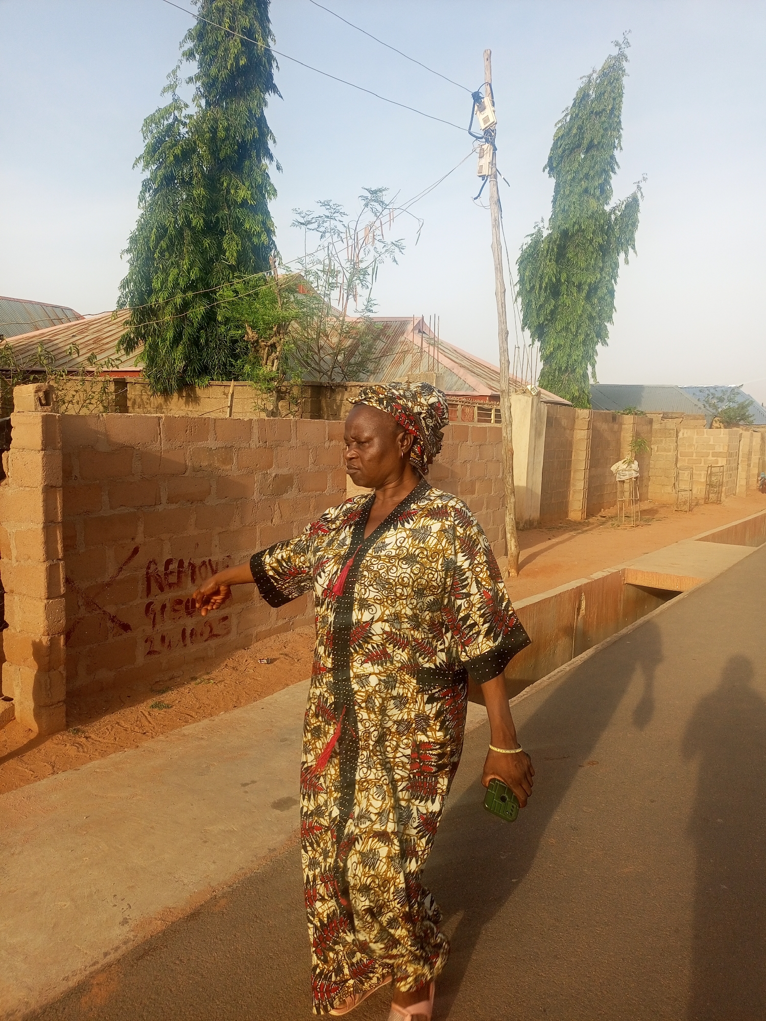 A woman wearing a patterned robe and head covering walks on a street next to a red brick wall marked with writing. She holds a green cell phone in her right hand, extending her left arm towards the wall. The surrounding environment includes tall trees, utility poles, houses with corrugated roofs, and a concrete drainage ditch along the street. The location is in a residential area, but the specific city and country are not discernible. The photograph is taken during daylight, possibly in the late afternoon, indicated by the long shadows.