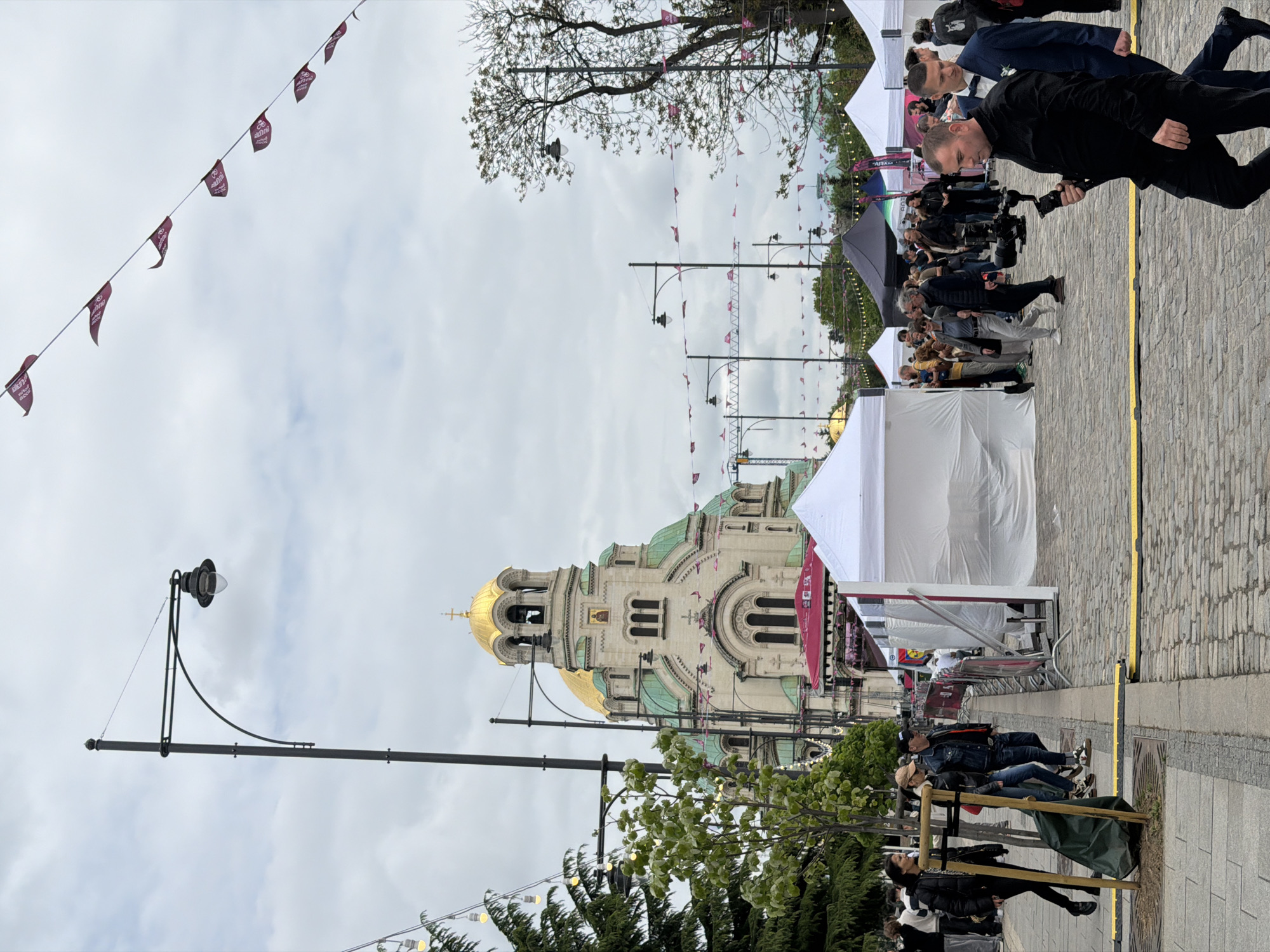 The image depicts a street scene in Sofia, Bulgaria, featuring the Alexander Nevsky Cathedral as a prominent landmark in the background. The sky is overcast. Streetlights are visible along the road. Pennant banners hang over the street. Tents are set up along the side of the road. People are seen walking and gathering along the street. Some people are walking with camera equipment. The street is paved with cobblestones. A yellow line is marked along the pavement.