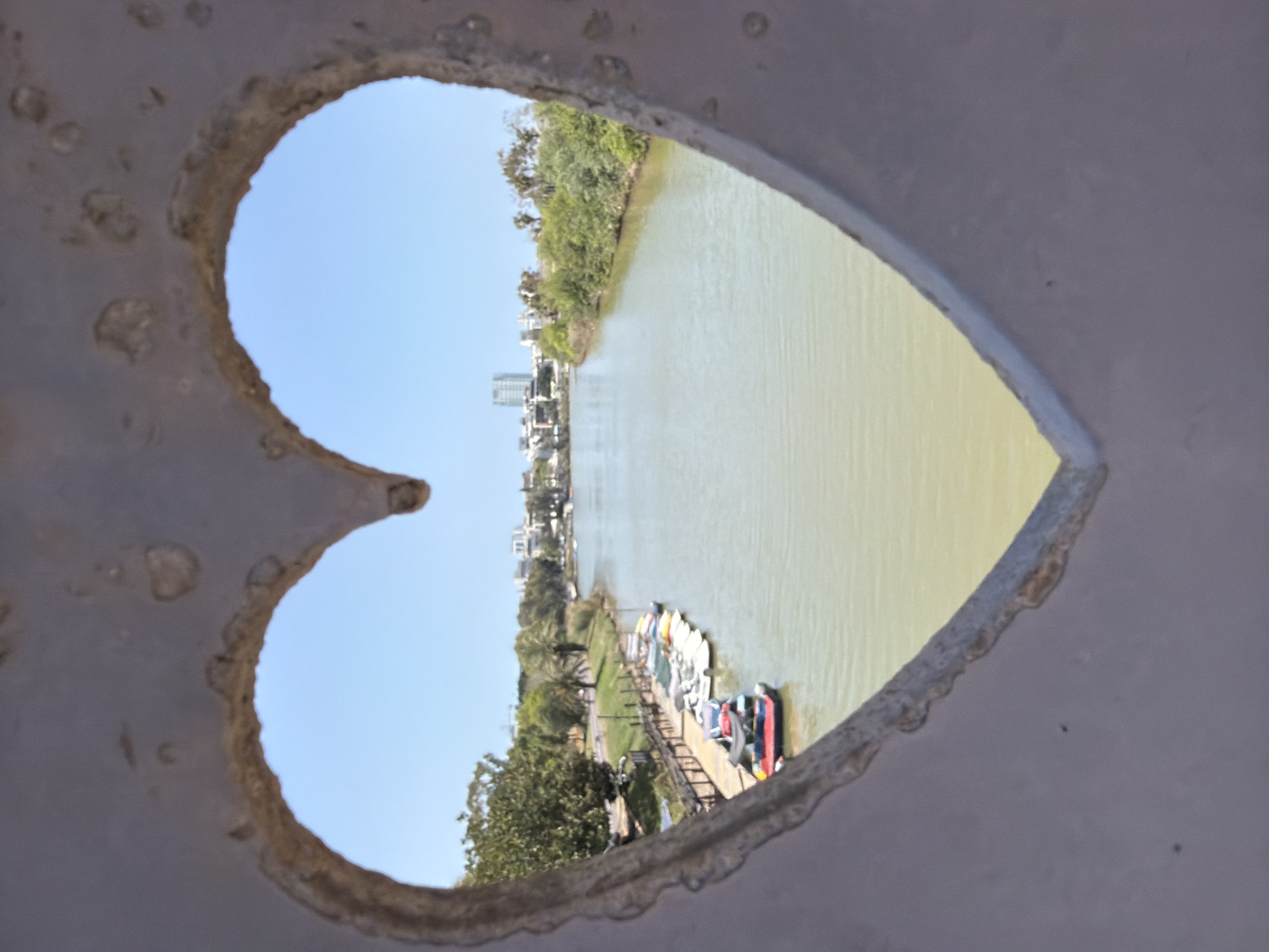 The image shows a view of the Yarkon River in Tel Aviv, Israel, as seen through a heart-shaped opening. The river is wide and the water is a pale green color. The banks of the river are lined with trees and greenery. In the distance, there are buildings and structures that suggest a city skyline. Nearer to the opening, there is a dock with boats. The opening is in a weathered, grey material with an uneven surface.