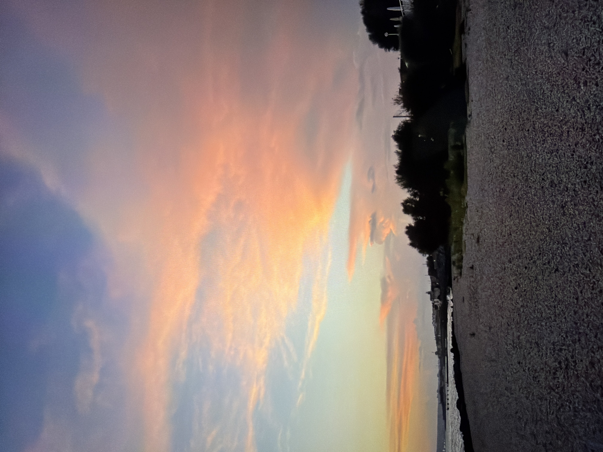The image displays a picturesque sunset or dusk scene over a body of water, likely in Ros Na Rí, Ireland. The sky, dominating the upper part of the frame, features a breathtaking display of colors, transitioning from soft blues and purples on the left to vivid pinks, oranges, and golden yellows towards the right, where the setting sun illuminates scattered clouds. These clouds range from wispy streaks to broader, diffuse formations.
In the midground, a dark, silhouetted line of dense trees and foliage stretches along what appears to be a bank or distant shoreline on the right side. Behind these trees, faint structures, possibly parts of buildings or railings, are barely discernible against the muted sky. A calm body of water, reflecting the soft light and subtle hues of the sky, occupies the central-left portion of the midground.
The foreground is composed of a dark, granular surface, characteristic of a gravel or pebbled path or beach, which runs horizontally along the edge of the water. No individuals or specific activities are present, allowing the natural beauty and tranquility of the twilight landscape to be the sole focus. The scene evokes a sense of peace and natural splendor at the close of day. There is no visible text in the image.