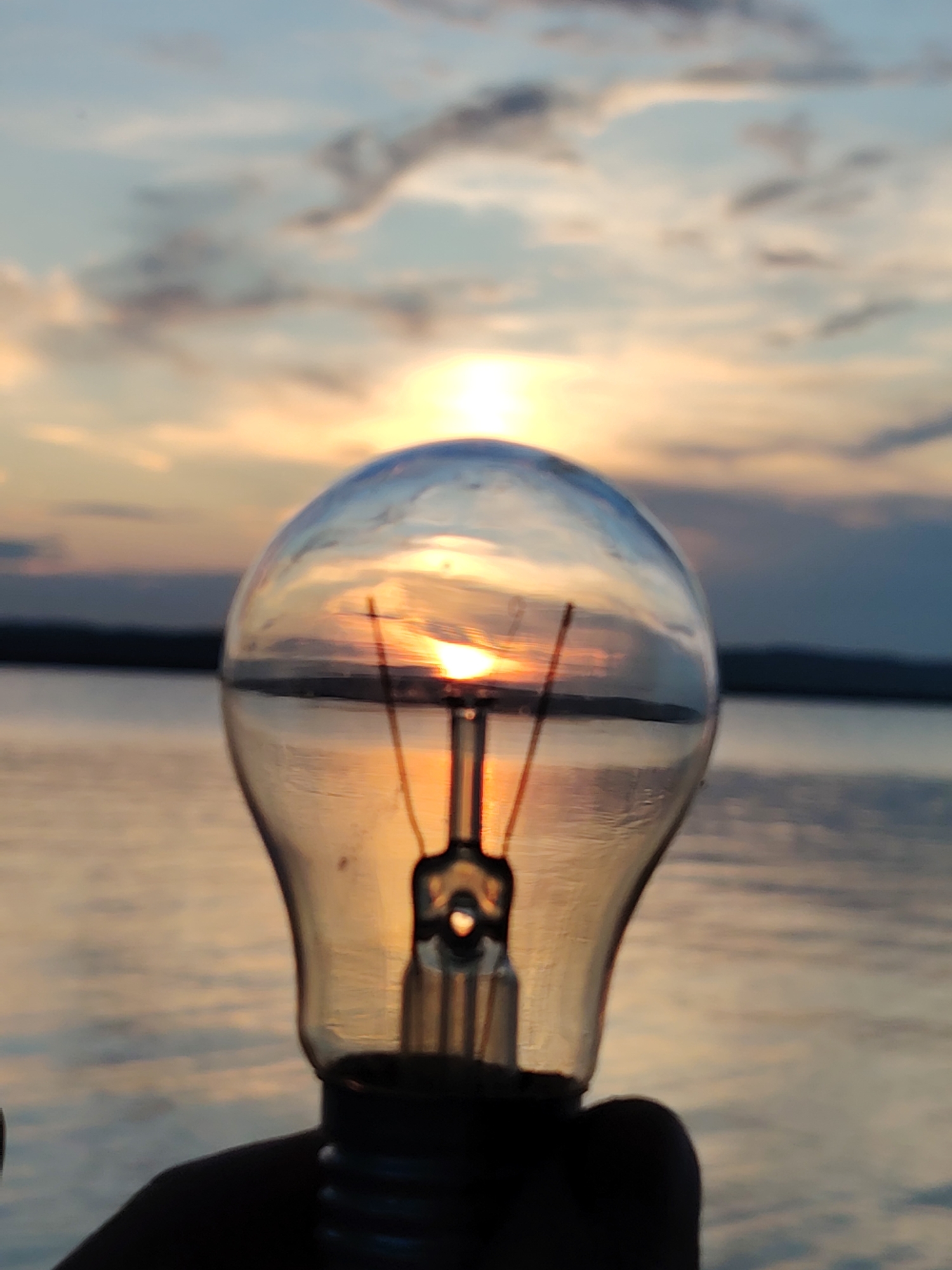 A close-up, slightly blurred shot features a clear glass light bulb held in silhouette against a sunset over a body of water. The sun is visible through the bulb, creating a lens effect, with its rays reflecting on the rippling surface of the water. The sky displays warm sunset colors, transitioning from orange and yellow near the horizon to pale blue and grey with wispy clouds higher up. In the background, a dark, low-lying landmass is visible on the horizon. The image appears to have been taken outdoors in an environment resembling a waterfront in the city of Chaikovsky, Russia, given the characteristic landscape of the Kama River or its reservoirs which are prominent features of the area.