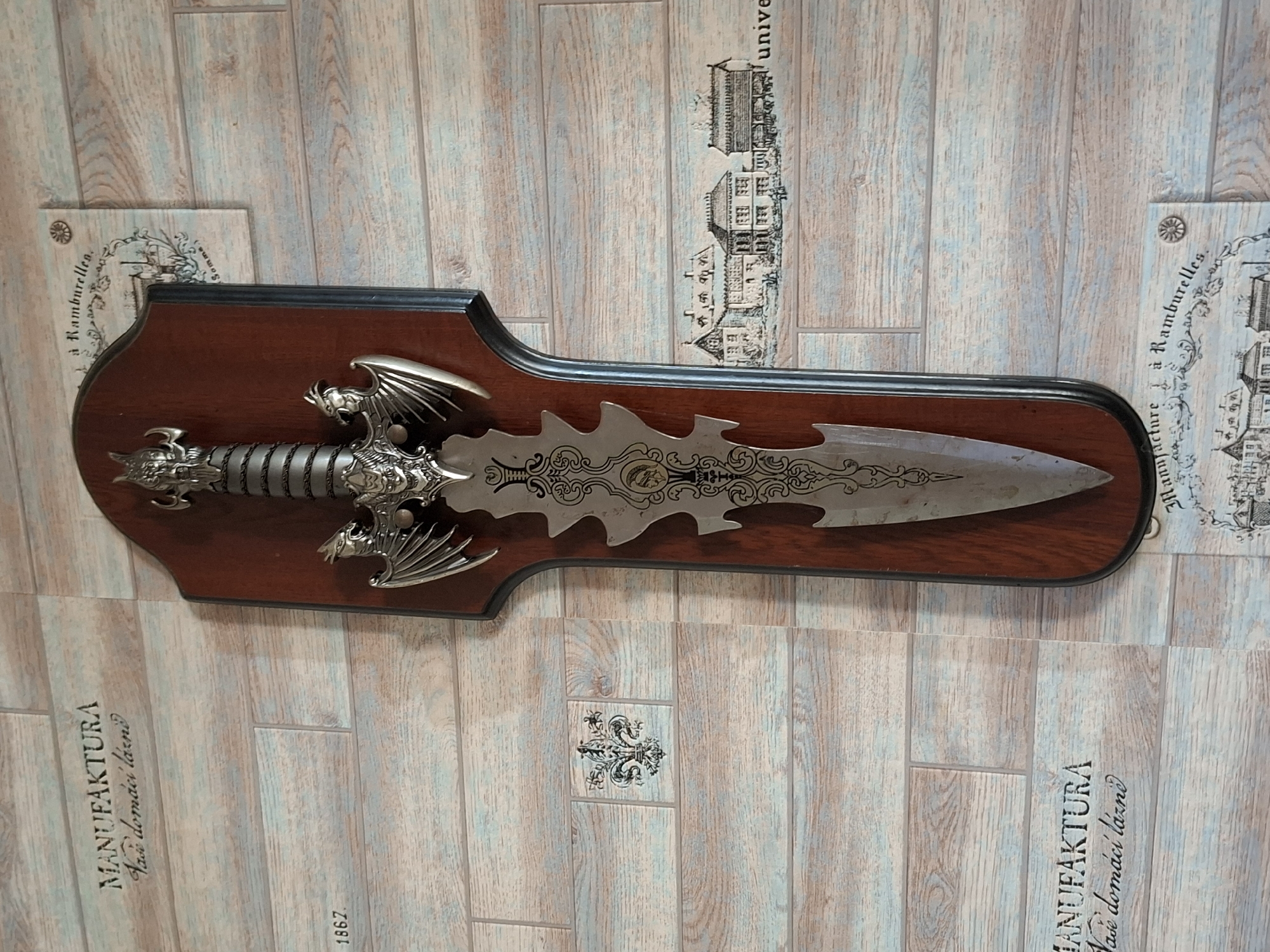 A decorative sword with a dragon motif is mounted on a wooden plaque against a wood-paneled wall. The wall features a pattern of horizontal planks with subtle blue and brown tones. Several small decorative prints are affixed to the wall. One print in the upper right corner depicts buildings and text. Another in the lower left corner contains the text "MANUFAKTURA Vase domáci lázně" and a fleur-de-lis emblem. A third print in the lower right corner shows more buildings and the word "unive". The sword itself has a metallic finish and is intricately detailed, with a dragon's head as the pommel and dragon wings on the hilt. The plaque is dark brown and has a sculpted edge. The overall scene is well-lit, and there are no people or animals visible. The location is not specified in the provided image or context.