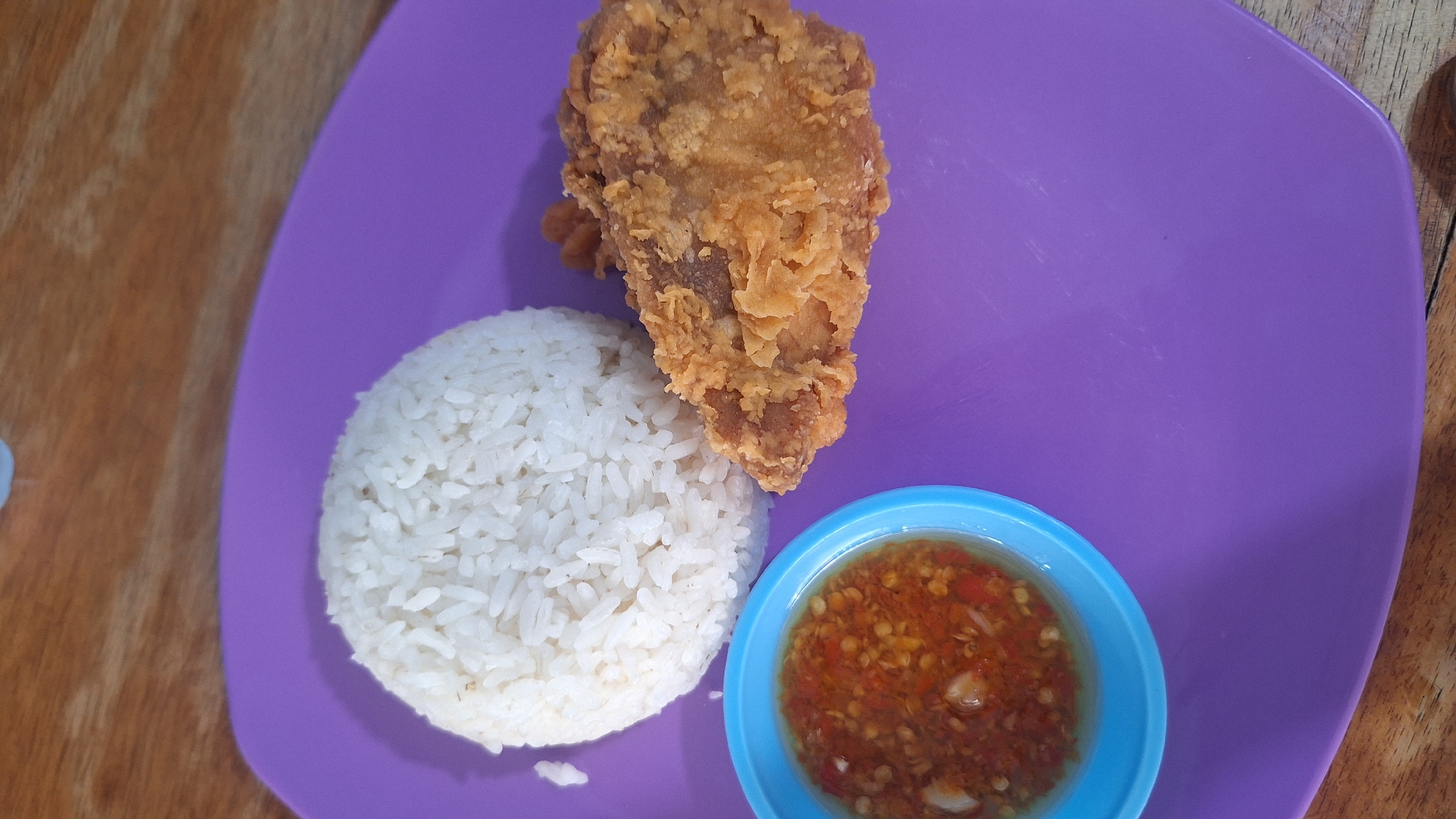 A plate of fried chicken, rice, and chili sauce sits on a purple plate. The plate rests on a wooden table. This photo was taken in Sukaharja, Indonesia.