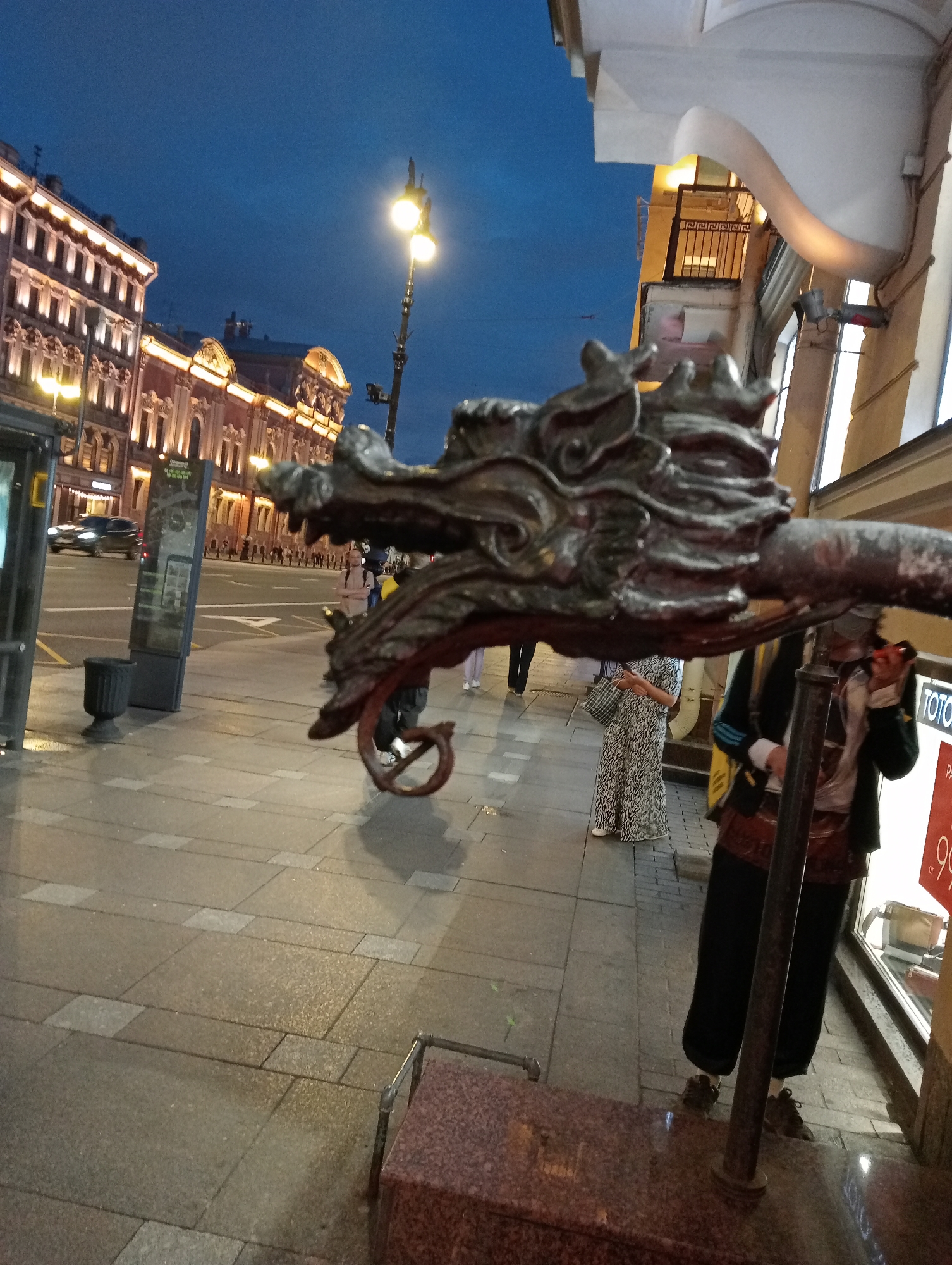 The image shows a close-up of a dragon-shaped sculpture in the foreground, with a street scene in Saint Petersburg, Russia, in the background. The sculpture appears to be made of dark metal and has intricate detailing. In the background, there are illuminated buildings with neoclassical architecture, a street with a few cars and pedestrians, and streetlights. The sky is dark, suggesting it is either dusk or nighttime. The overall impression is a blend of historical architecture and urban life.