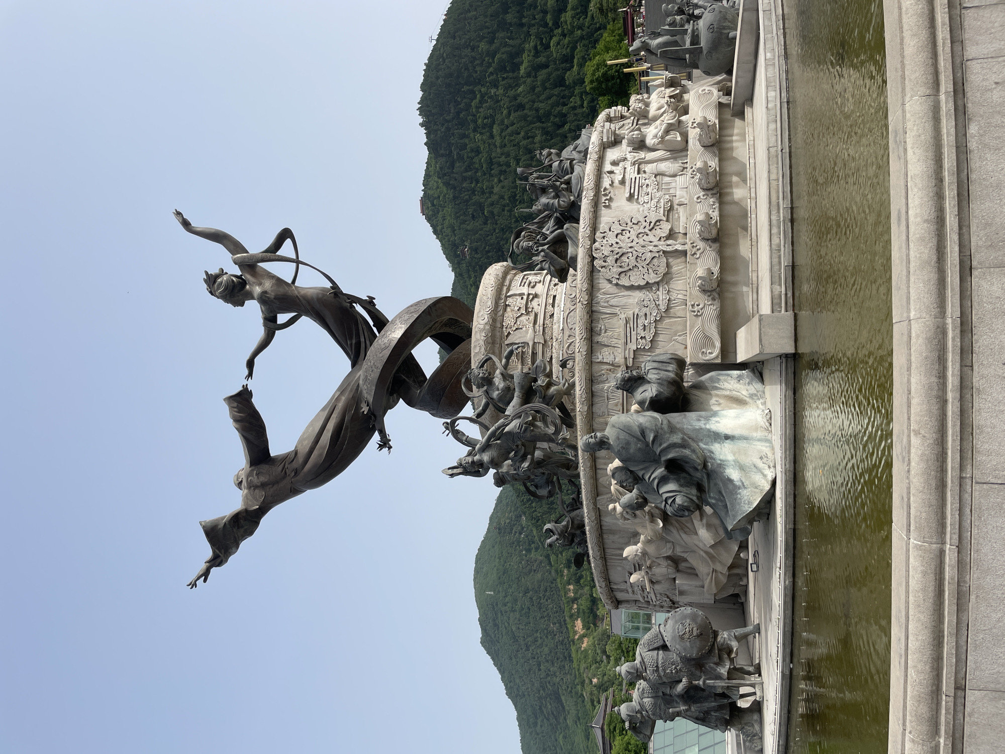 The image depicts a large, ornate sculpture in Lintong District, China, set against a backdrop of a green, mountainous landscape and a pale blue sky. The sculpture is primarily composed of bronze and stone elements. The upper portion features a bronze sculpture of two figures in flight, one partially draped in flowing robes and the other adorned with a headpiece. Below these figures, a large, decorative stone relief structure with intricate carvings and a bas-relief of various figures is evident. Additional bronze sculptures of people in traditional garb encircle the base of the structure. A pool of water surrounds the base of the sculpture, adding a reflective element to the scene. The composition appears to be photographed from a low angle.