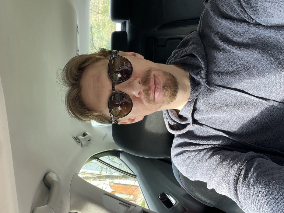 A male subject is seated in the rear passenger compartment of a vehicle in Monsummano Terme, Italy. He has light brown hair, a goatee, and a mustache. He wears round, dark-lensed sunglasses with a dark, possibly tortoise-shell, frame. He is dressed in a textured, dark grey hoodie. Sunlight illuminates his left side, casting shadows. The vehicle's interior includes a light grey headliner and dark grey/black seating and door panels. Through the vehicle's windows, green foliage from trees and a portion of a light-colored building with an arched opening are visible. Reflections in the sunglasses indicate the presence of a camera device, likely held by the subject. The man maintains a neutral expression.