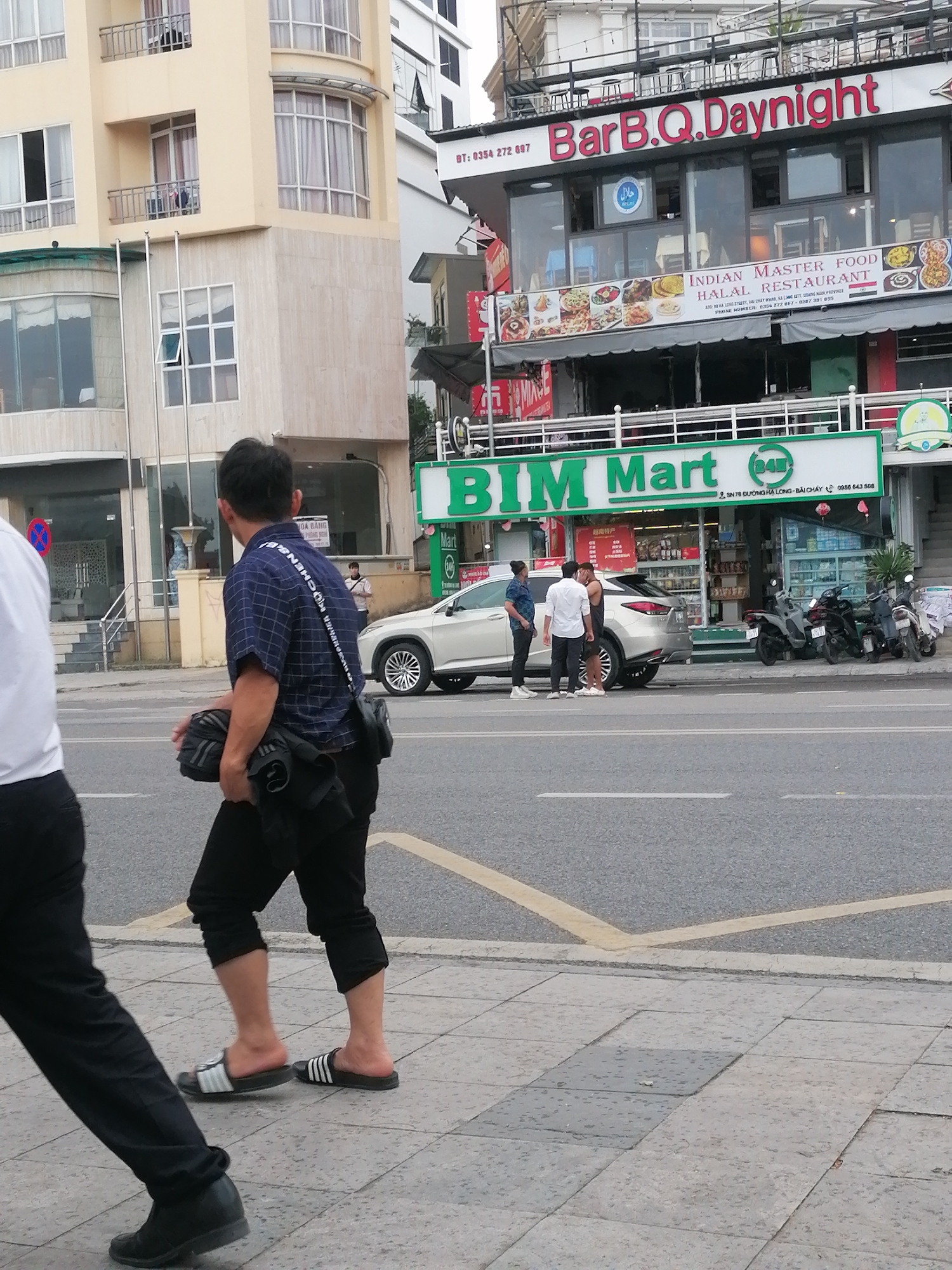 The image shows a street scene in Bai Chay Ward, Vietnam. A man in a blue plaid shirt, dark cropped pants, and Adidas sandals walks on the sidewalk toward the left side of the image. A person partially obscured in white clothing is walking to the left of him. Across the street is a "BIM Mart" grocery store, in front of which several people are standing next to a white car. Three motorbikes are parked nearby. Above the grocery store is the "BarB.Q.Daynight" restaurant, advertising Indian Master Food. The scene includes buildings, sidewalks, streets, vehicles, and pedestrians.