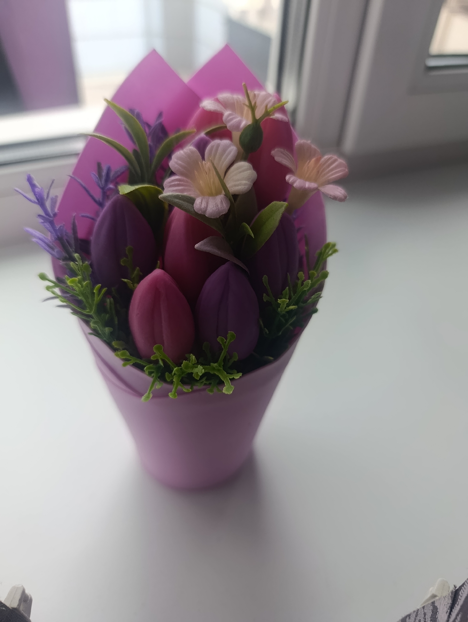 A bouquet of artificial tulips and lavender is displayed in a purple pot on a white surface, possibly a windowsill, with a blurred background suggesting an indoor environment with a window. The tulips are predominantly purple and pink, with a few small light pink flowers interspersed. Green foliage and sprigs of artificial lavender add to the floral arrangement. The pot is wrapped in pink paper. The scene is illuminated by natural light coming from the side. There are no people or animals visible in the image. The overall impression is that of a decorative item.