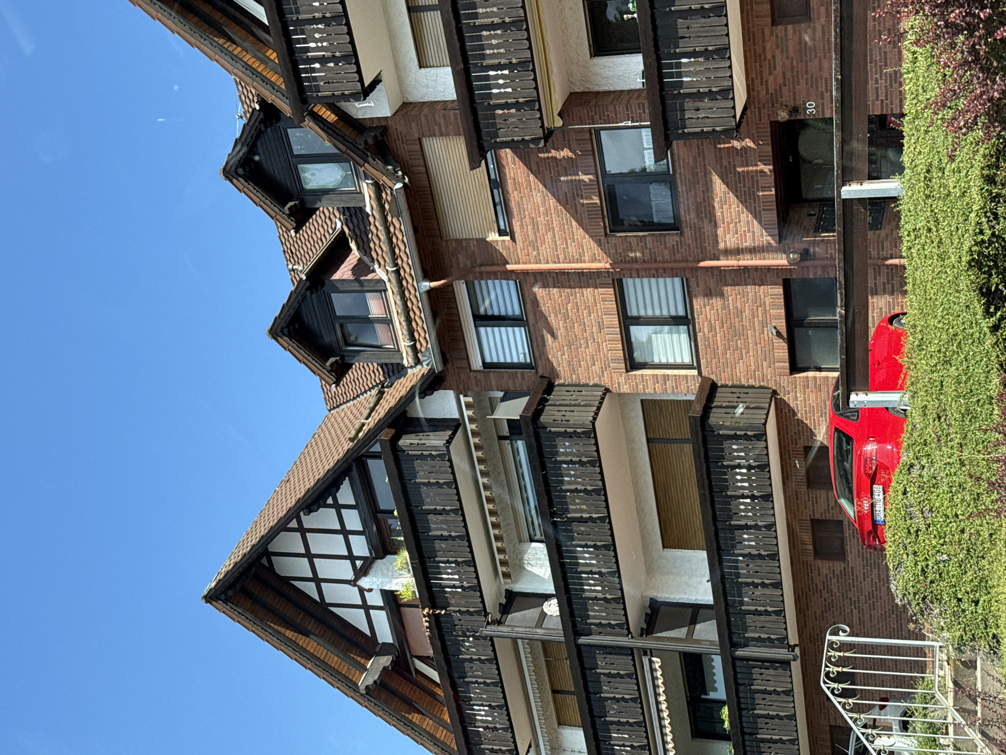 The image captures a multi-story building with a distinct architectural style, likely residential, located in Bad Sachsa, Germany. The building features red brick lower levels and upper levels with white stucco and dark wooden balconies and eaves. The roof is tiled and has several dormer windows. A bright red sedan is parked in front of the building, partially obscured by lush green bushes. A white metal railing and stairs lead up to the building. The sky above is clear and blue, indicating a sunny day. The building displays the number "30" above an entrance. There are no people visible in the image.