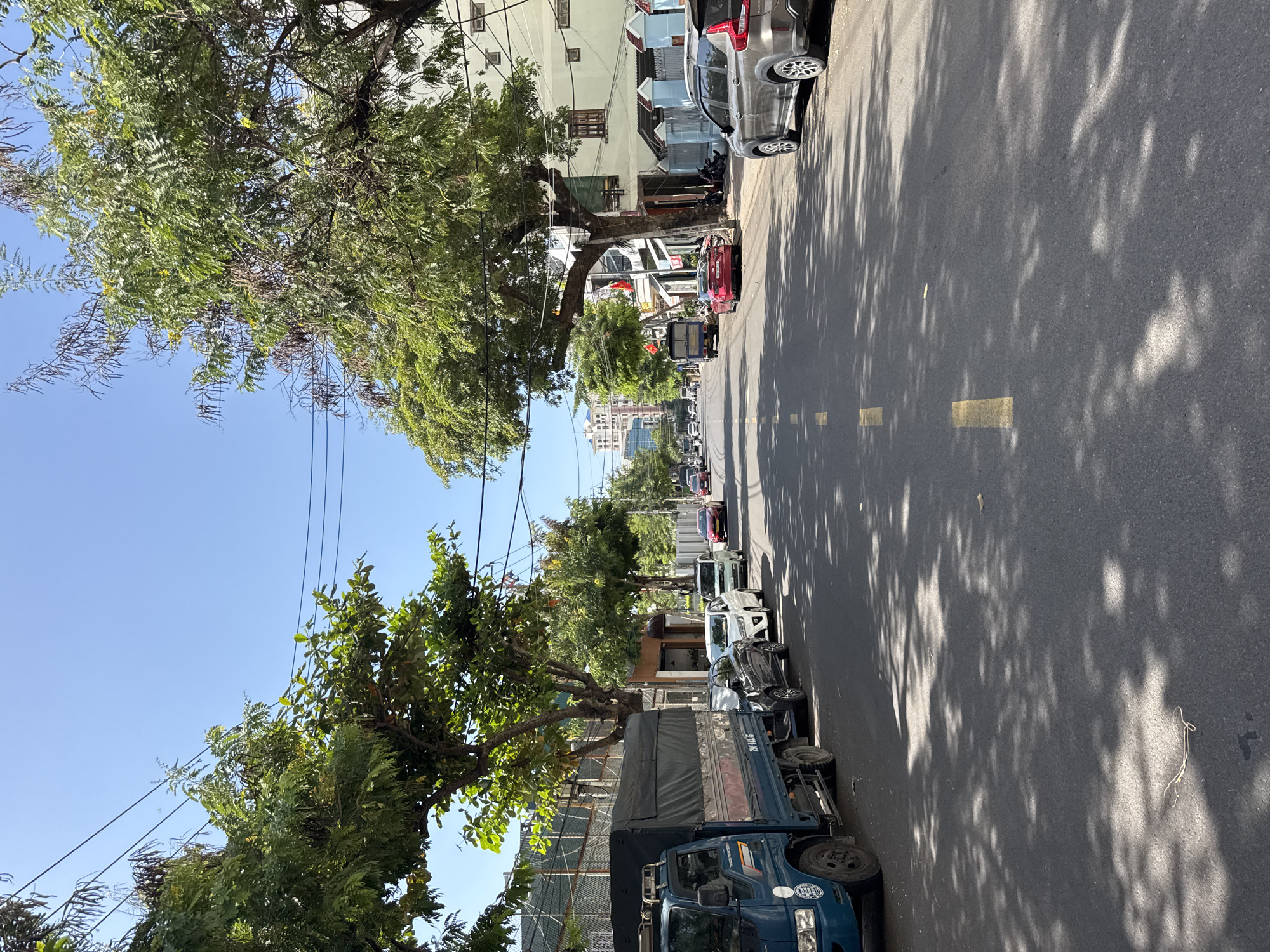 The image depicts a street scene in Nha Trang, Vietnam, on a bright, sunny day. A blue truck with a black tarp-covered cargo bed is parked on the left side of the street. Several cars are parked along the right side of the road, including a dark sedan, a white sedan, a white SUV, a red sedan, and a silver SUV at the far right. The street has yellow lane markings. Lush green trees line both sides of the street, casting dappled shadows on the asphalt. Buildings, some with balconies and windows, are visible behind the trees. In the distance, taller buildings can be seen. The sky is clear and blue. There are no people clearly visible or engaged in activity.