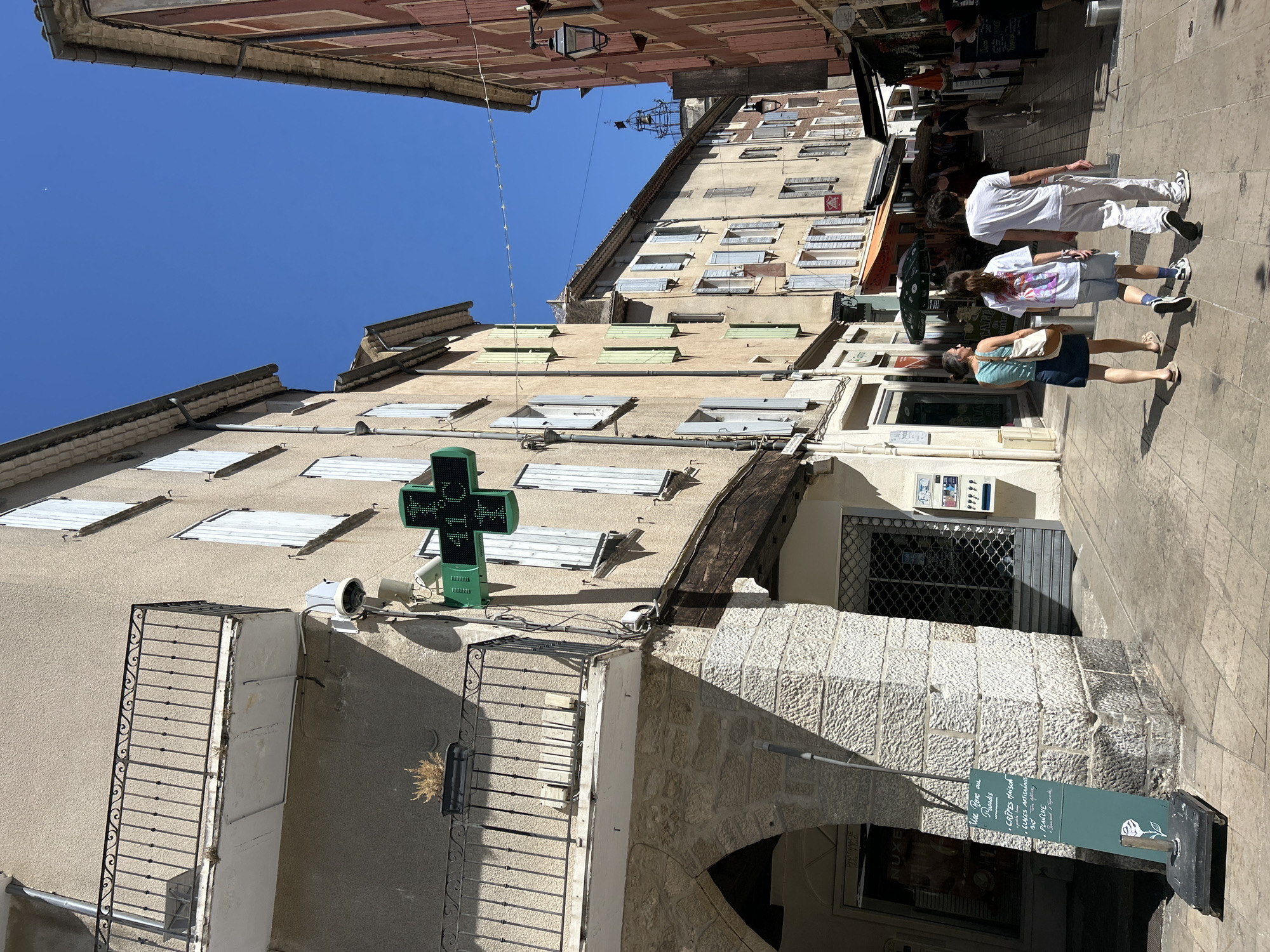 The image shows a street scene in Nyons, France, on a sunny day. A narrow street is lined with buildings, some with light-colored facades and blue shutters, others with pinkish-red facades. There is a prominent green pharmacy sign with a cross symbol and the temperature displayed as 41°C.
In the foreground, three people are walking away from the camera down the street. The person on the left is a woman wearing a striped tank top, a blue denim skirt, and sandals, carrying a beige bag. The person in the middle is a younger person wearing a white t-shirt with a graphic print, denim shorts, and white sneakers. The person on the right is a man wearing a white t-shirt and white pants with white sneakers.
To the left of the street, a stone archway leads to an entrance. A green sign with white text and a white rose graphic is propped up near the archway. Above the archway, a balcony with a black wrought-iron railing is visible.
Further down the street, awnings and umbrellas suggest the presence of shops and cafes. In the distance, a bell tower can be seen above the rooftops. The sky is a clear, bright blue.