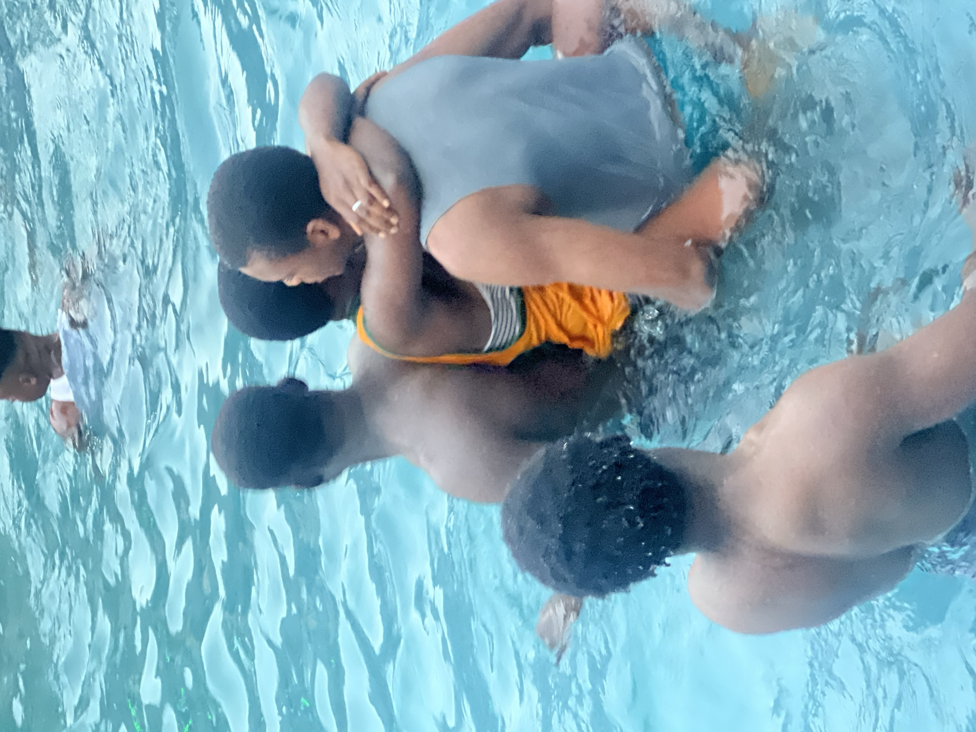 This image, taken in Nkpor, Nigeria, captures a vibrant scene of several people engaged in playful activity within a clear, light blue swimming pool, viewed from an overhead perspective.
The central focus of the photograph is on two individuals who appear to be embracing or playfully wrestling in the water. One person, dressed in a grey t-shirt and blue or teal swim shorts, is positioned behind the other, with their arms wrapped around them. A silver ring is noticeable on the left hand of this individual. The person they are holding wears an orange or yellow sleeveless top featuring vertical stripes. Their faces are largely obscured or turned away from the camera, but their body language suggests a joyful and recreational interaction.
Around these two central figures, other people are partially visible, contributing to the lively atmosphere. To the left side of the frame, the head and shoulders of an individual wearing a white shirt can be seen emerging from the water. In the bottom right corner, the back and dark, curly hair of another person are visible as they navigate the pool. Another person's back and the top of their head are partly submerged towards the bottom left of the image.
The water exhibits dynamic ripples and splashes, indicative of movement and active play. The overall lighting suggests it is daytime. The scene portrays a communal and enjoyable gathering, likely an informal social event or leisure activity in the swimming pool. No discernible text is visible within the image.