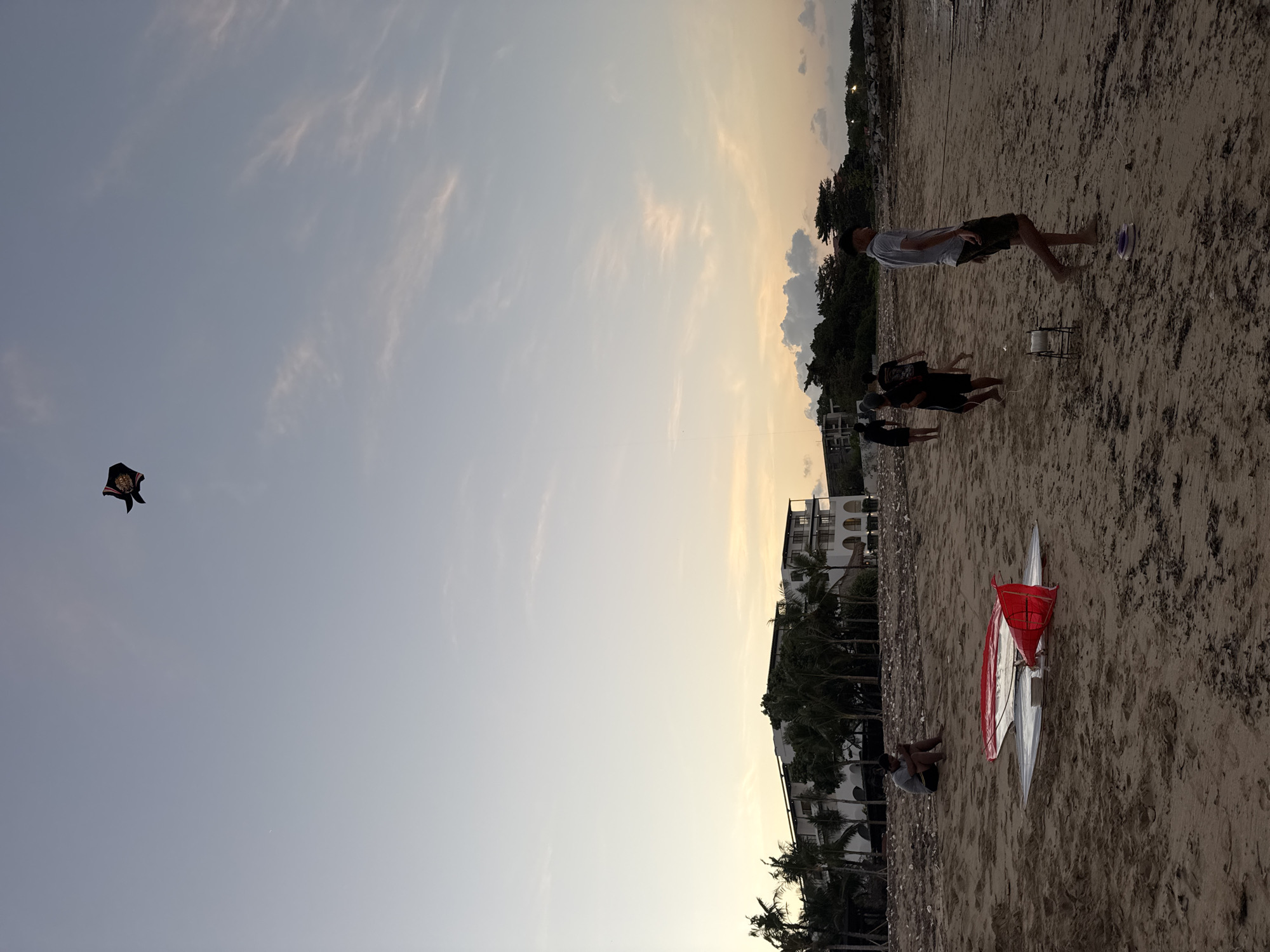 A group of people are on a sandy beach in Kuta Selatan, Indonesia, with a kite flying in the sky. The sky is partly cloudy with a gradient of soft blues and yellows, suggesting it is either dawn or dusk. In the background, a modern white hotel with balconies and palm trees is visible. On the beach, several individuals are walking or standing, some appearing to be engaged in flying the kite. A red and silver kite lies on the sand in the foreground, alongside a spool of kite string. One person is seated on the beach near the kite. The sand is a light brown color and appears to have some debris scattered across it. The overall atmosphere is relaxed and recreational.