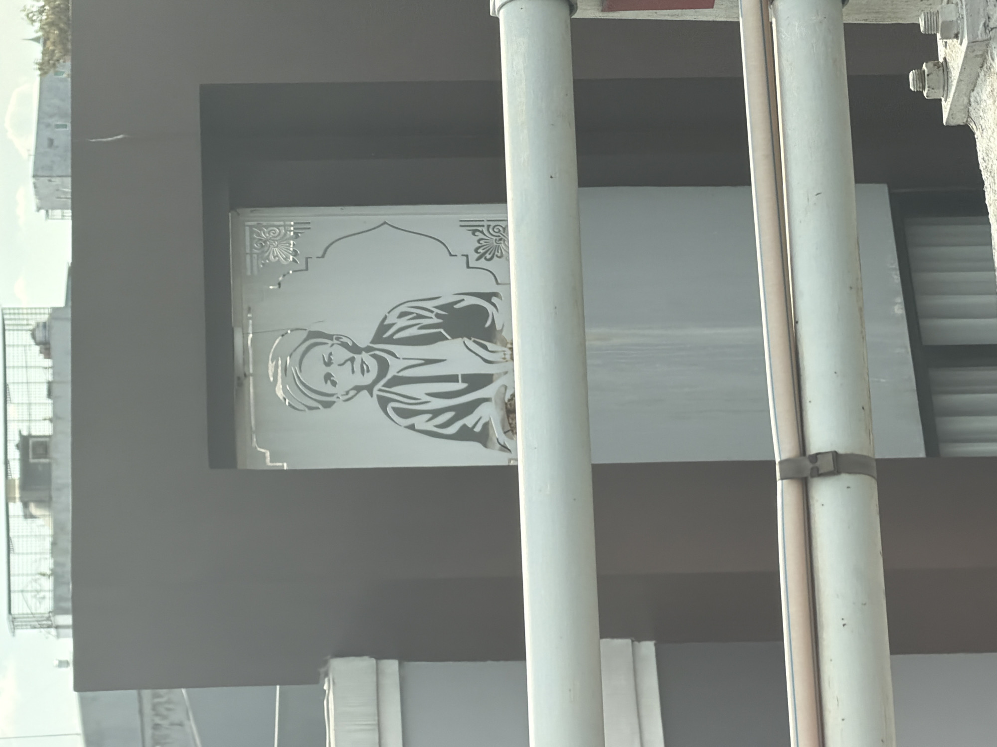 The image shows a graphic depiction of a man on a sign, positioned within a dark gray rectangular frame attached to a building exterior. The backdrop behind the sign appears to be an open space, possibly a window, allowing visibility to another building in the distance. Two vertical white pipes are positioned in the foreground, partially obstructing the view of the sign. The pipes are fastened together with a metal clip. Based on prior information, the location of the image is likely within North Jakarta, Indonesia.