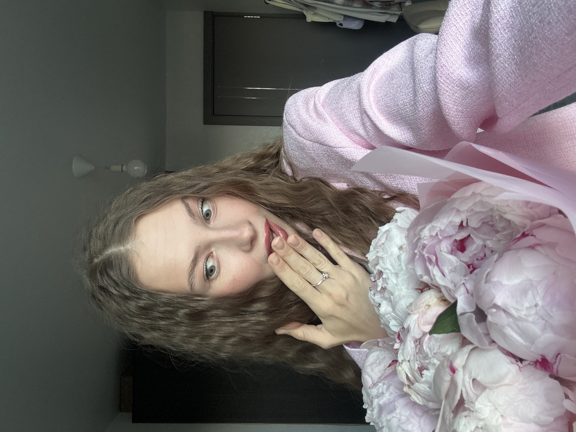 The media file is a portrait of a young woman with long, curly brown hair and light green eyes. She is wearing a pink tweed jacket and holding a bouquet of pale pink and white peonies. Her left hand is raised to her face, touching her cheek, and she is wearing a silver ring on her right ring finger. Her expression is one of surprise or excitement, with her mouth slightly open and her eyes wide. The background is a dimly lit room with a grey wall, a light fixture, and a dark doorway or closet. The lighting suggests it could be daytime or evening indoors. The overall mood is cheerful and intimate, possibly capturing a moment of receiving a gift. There is no visible text in the image. The location context is Perm, Russia.
