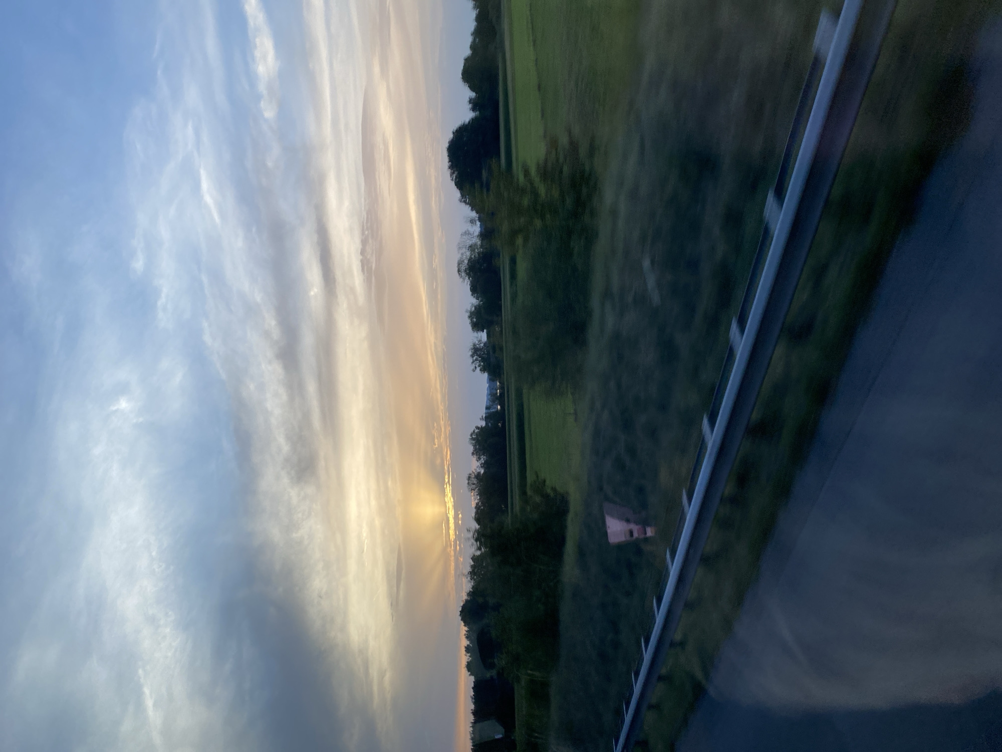 The image depicts a sunset over a rural landscape, likely in the vicinity of Chiemsee, Germany, as indicated by the context. The sky is a blend of blues, grays, and soft oranges and yellows, with the sun's rays fanning out from behind a band of clouds near the horizon. Below the sky, a line of trees borders a vast green field. In the foreground, a dark asphalt road is visible, separated from the field by a metal guardrail. The overall impression is of a serene, natural setting during twilight.