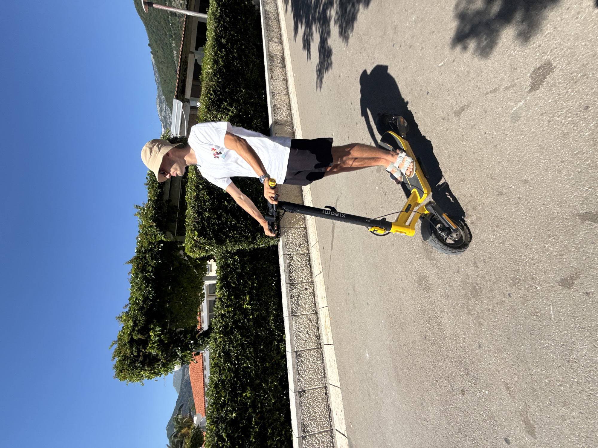 A person is riding a yellow Xiaomi electric scooter on an asphalt road. They are wearing a white t-shirt with a graphic, black shorts, a beige bucket hat, sunglasses, and white sandals. The scooter has a black handlebar and stem with yellow accents. The person is casting a shadow on the road. The background consists of a manicured hedge, a stone wall, and buildings with red-tiled roofs, with mountains visible in the distance under a clear blue sky. The location is the city of Budva, Montenegro.