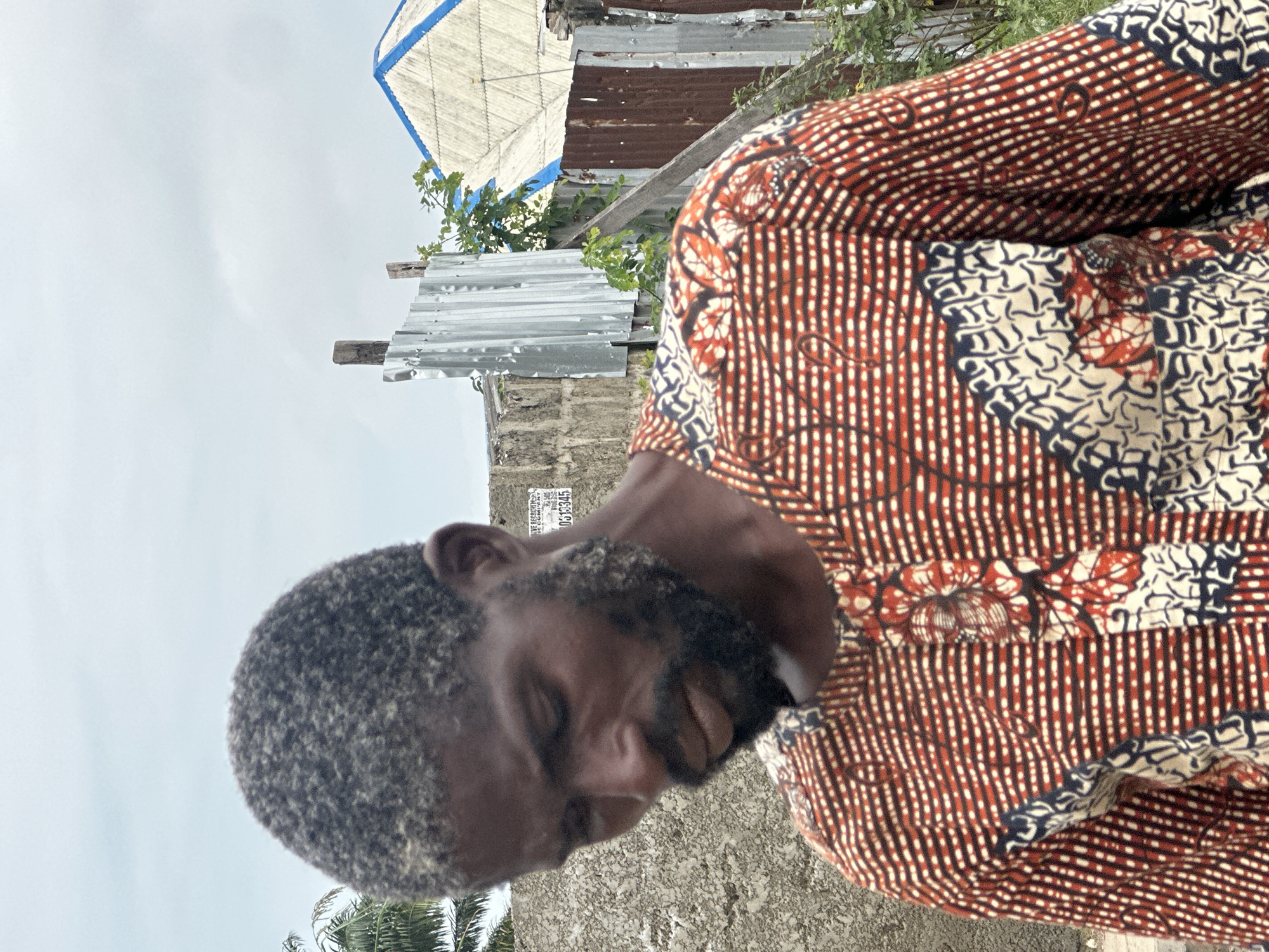 A medium shot captures a Black man in Badagry, Nigeria, from the chest up, looking down. He has short, graying hair and a full beard. He wears a vibrantly patterned shirt with red, white, and black stripes and floral motifs. Behind him, a weathered concrete wall is visible, with a faded sign bearing text that is mostly illegible but includes "053345". Further in the background, corrugated metal fencing and structures, including a roof with blue trim, suggest an outdoor, potentially informal setting. The sky above is overcast.