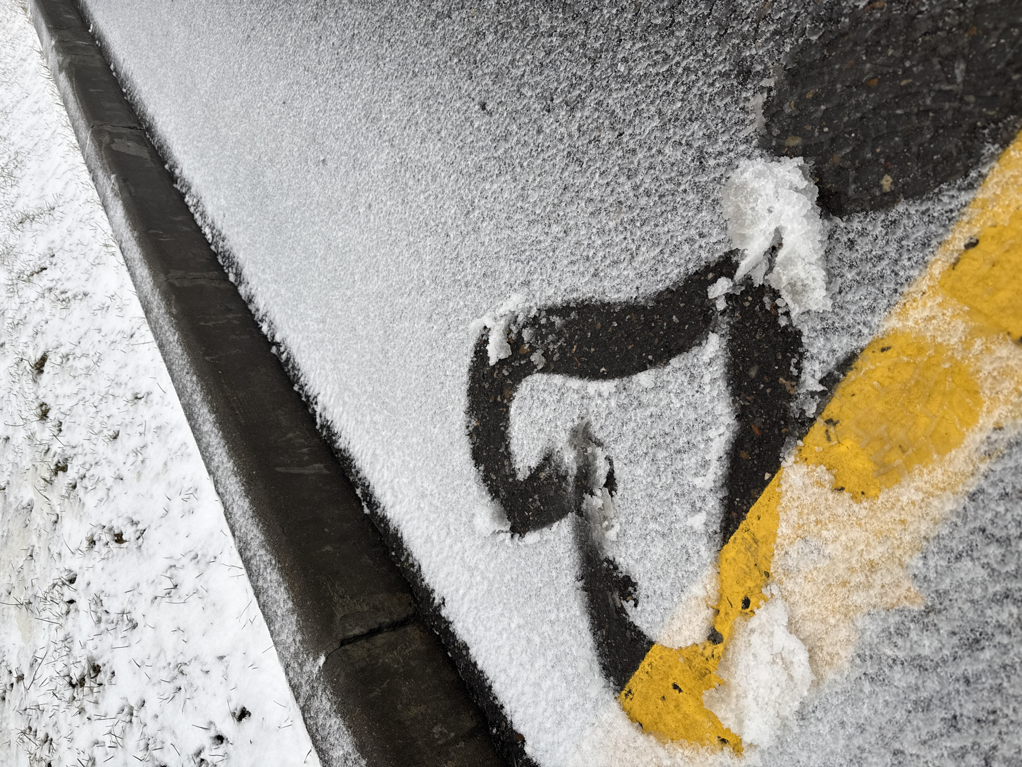 The image shows a section of asphalt road in Lupanovo, Russia, partially covered in a layer of snow or ice. A grey concrete curb runs along the left side of the image. A yellow road marking is visible on the asphalt surface. The snow is concentrated on the asphalt and partly covering the road marking.