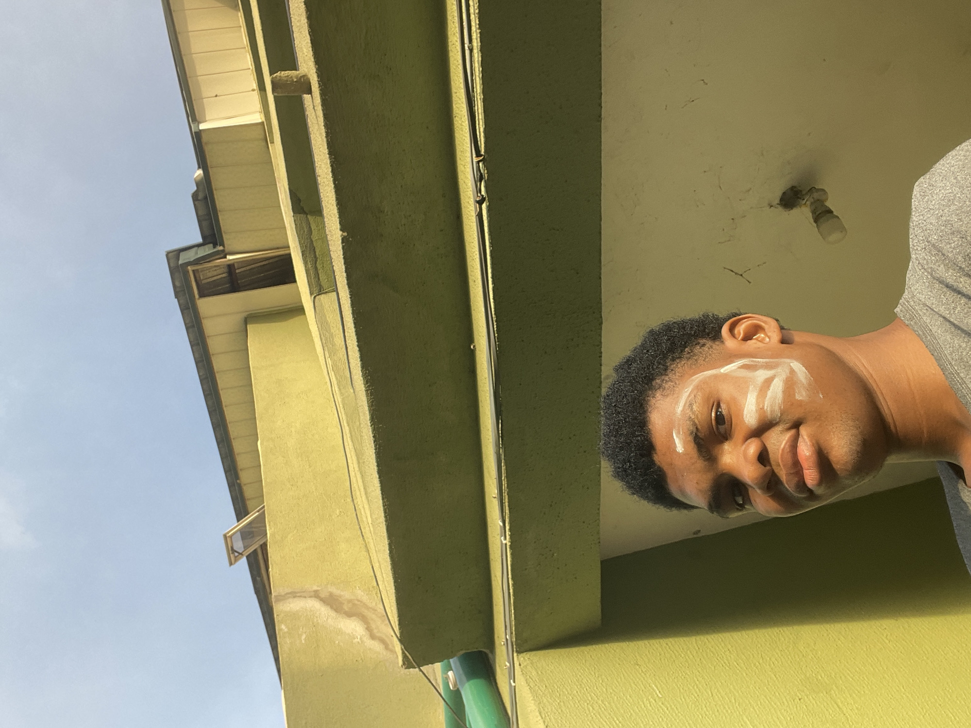 An eye-level, slightly tilted shot of a person with dark hair and skin stands under the eave of a building, looking into the camera. Patches of white cream are on their face. The building has a green exterior, and exposed wiring runs along the eave. The sky is visible in the background. The location is likely a residential area in Ado, Nigeria.