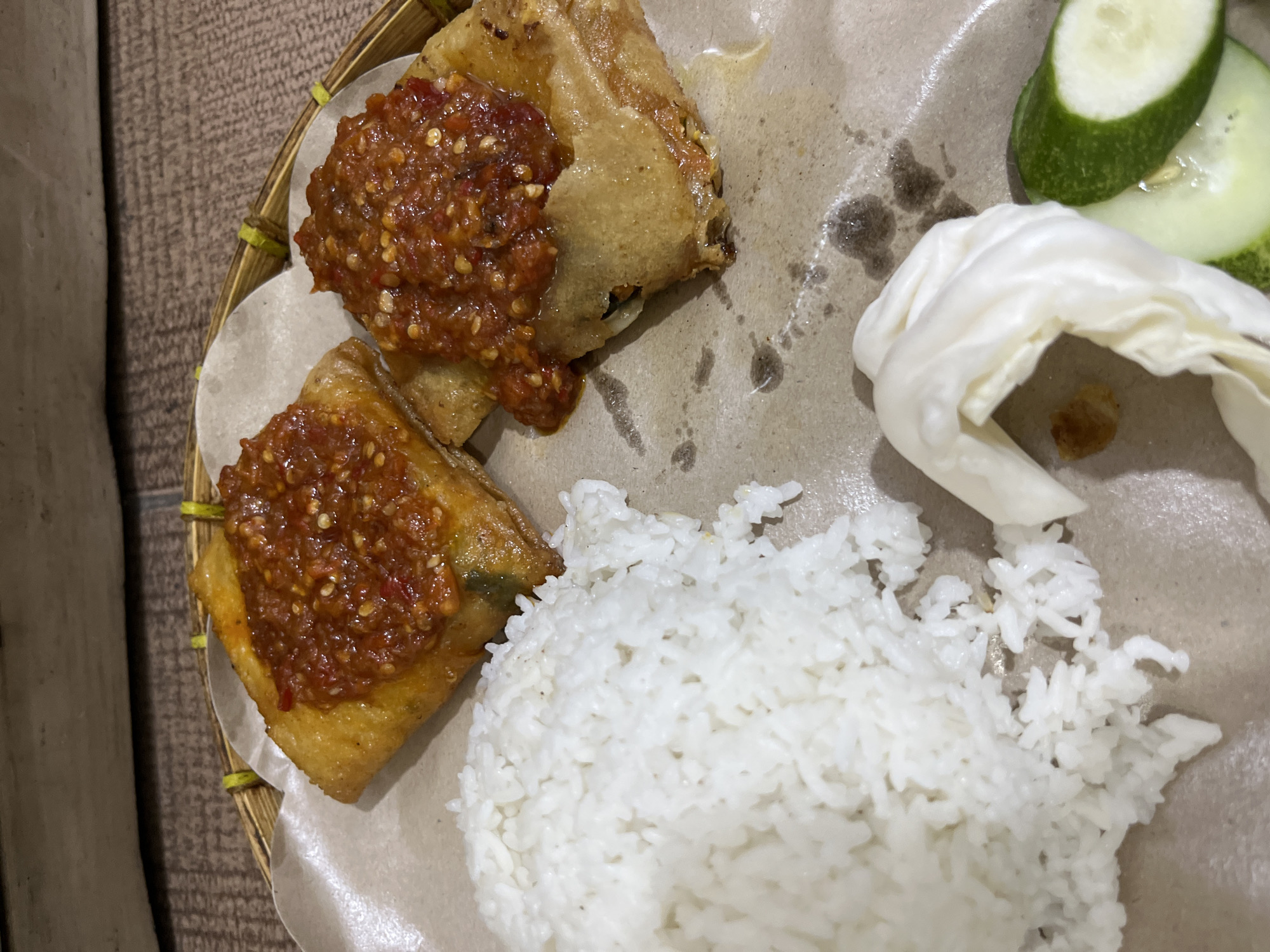 In a close-up, overhead shot, a plate of food is presented on a woven bamboo tray. The plate holds a portion of white rice, two pieces of golden-brown fried food topped with a red chili sauce, a few slices of cucumber, and a small pile of shredded white cabbage. The fried items appear to be savory snacks, possibly Murtabak or Martabak Telur, given their square shape and filling. The chili sauce is chunky with visible pieces of chili and other ingredients. The food is served on brown paper liners within the bamboo tray. The background consists of a textured surface, possibly a wooden table. The lighting is dim, suggesting an indoor setting, potentially a restaurant or home. The image does not contain any people or indicate a specific location in Central Jakarta, Indonesia.