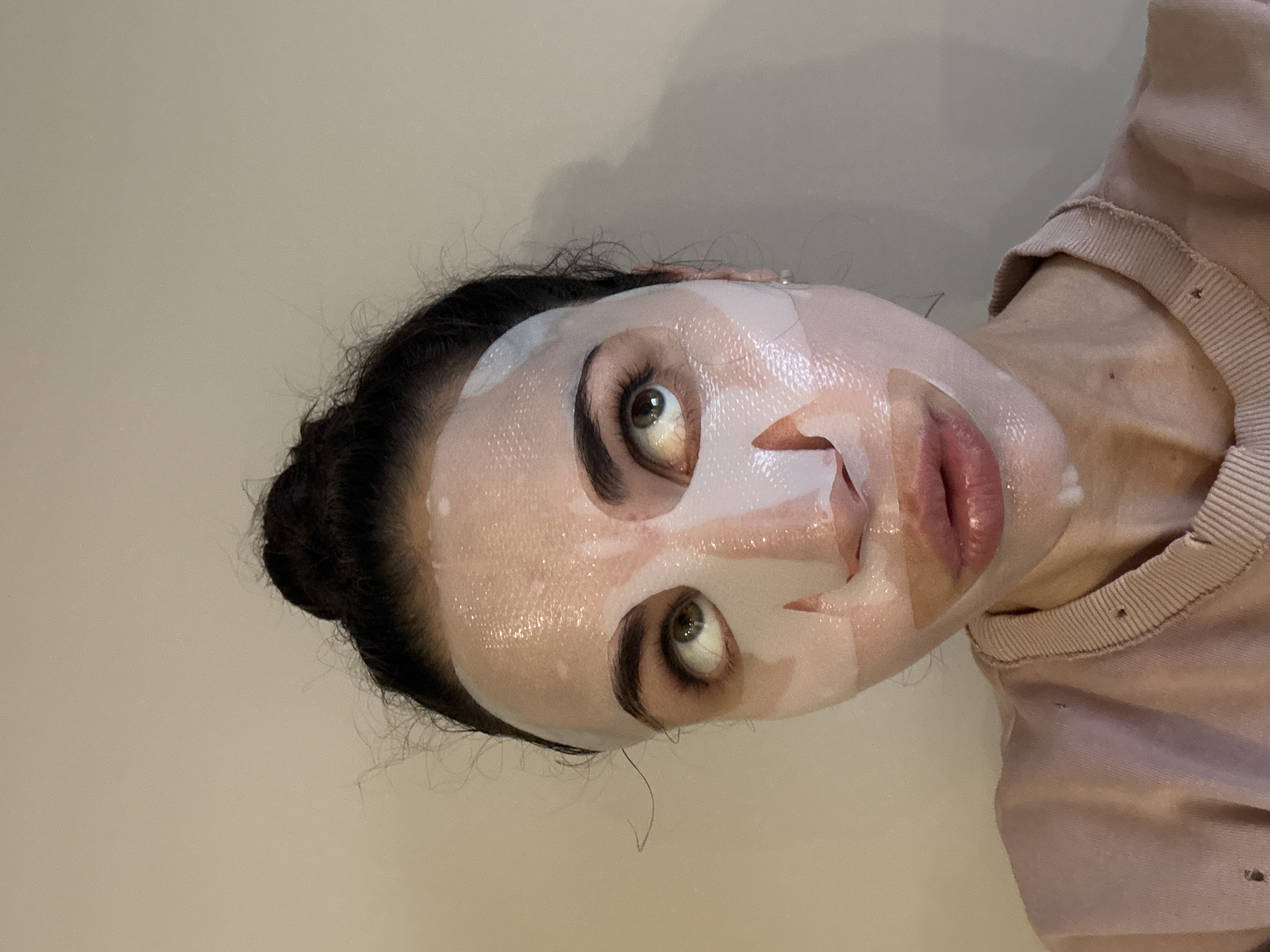 A woman with her hair in a bun is wearing a sheet face mask. She is wearing a pink shirt with small holes on the shoulder. The background is a plain wall. The location is not identifiable from the image.