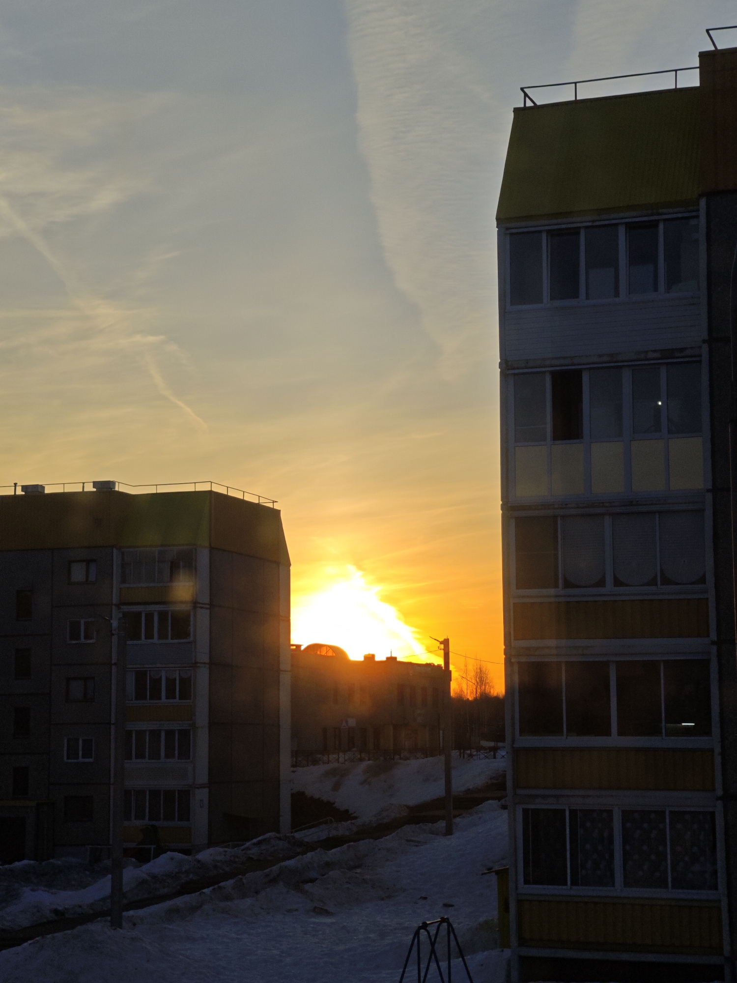 The image shows a winter sunset in Марковский, Russia. Two multi-story apartment buildings frame the scene, with the setting sun visible between them. Snow covers the ground. The building on the right is closer and partially obscures the view, while the building on the left is more distant. The sky displays streaks of pale clouds.