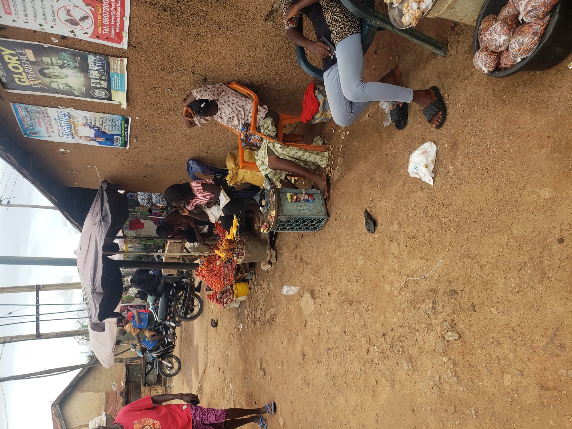 The image depicts an outdoor market scene in Television, Nigeria, with a focus on several individuals selling produce and other goods from stalls. In the foreground, a person wearing a red t-shirt and patterned shorts walks away from the camera, their blue flip-flops visible. To their left, a motorcycle carrying a rider and passenger is visible. Further into the scene, several stalls are set up, displaying piles of tomatoes, corn, and other food items. Three women are seated on chairs at a stall, with one appearing to be reading a book. A fourth person, wearing a leopard-print top and light gray leggings, is seated on a chair on the right side of the frame, facing away from the camera. A dark-colored bucket filled with goods wrapped in plastic bags is also visible in the foreground. The ground is dirt and sandy, with scattered debris. The background includes buildings with corrugated roofs and posters displayed on a wall. The overall atmosphere suggests a busy marketplace in a somewhat arid environment.