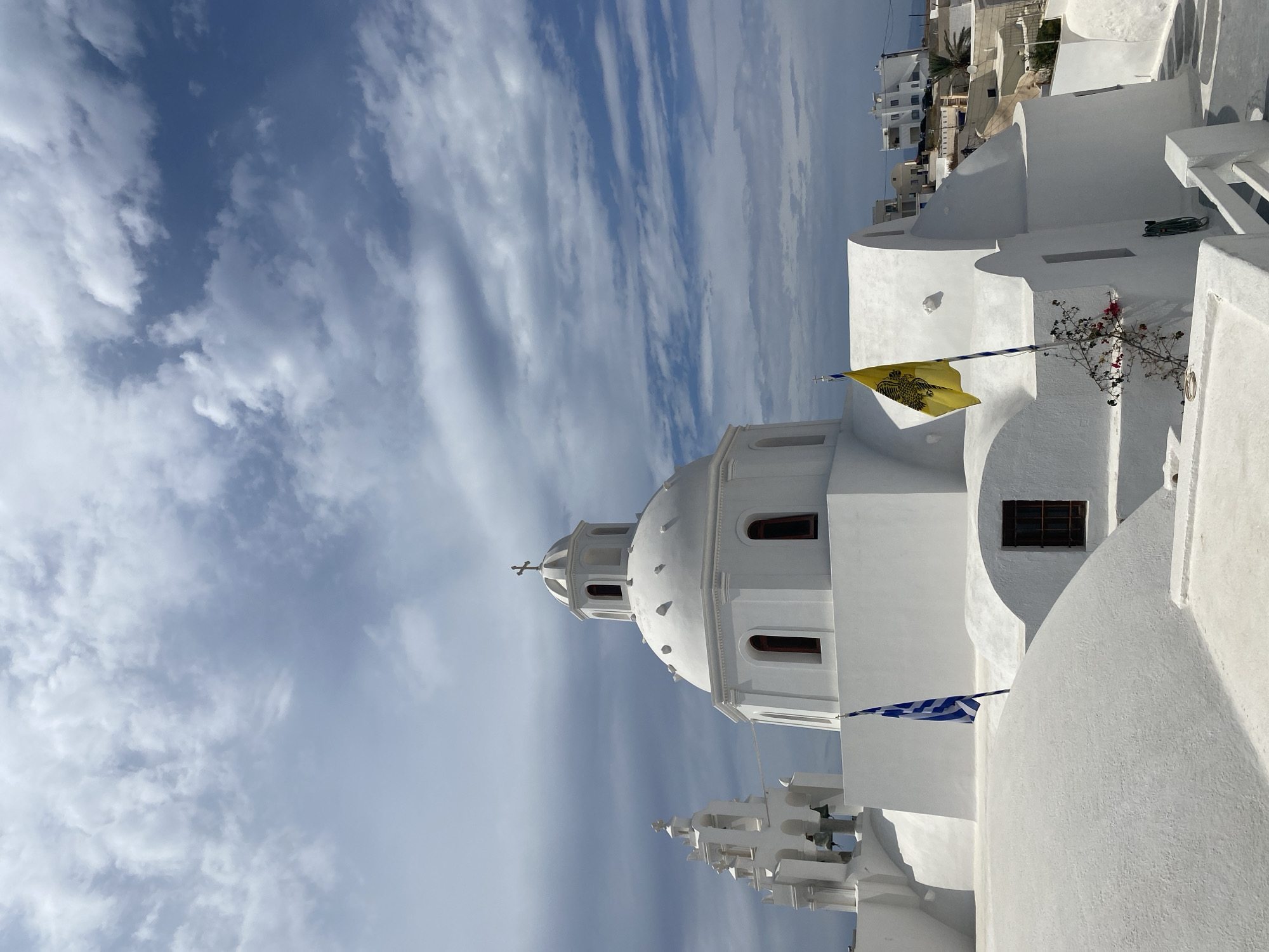 This is a high-angle, full shot of a white building with a domed roof in the city of Thira Municipal Unit, Greece. The building is a church, and it has a bell tower on the left. There are two flags flying in front of the church, one Greek flag and one yellow flag with a black eagle. The sky is cloudy and blue. The building is made of white stucco and has red-trimmed windows. There are steps leading up to the building. In the background, there are other white buildings and some palm trees.