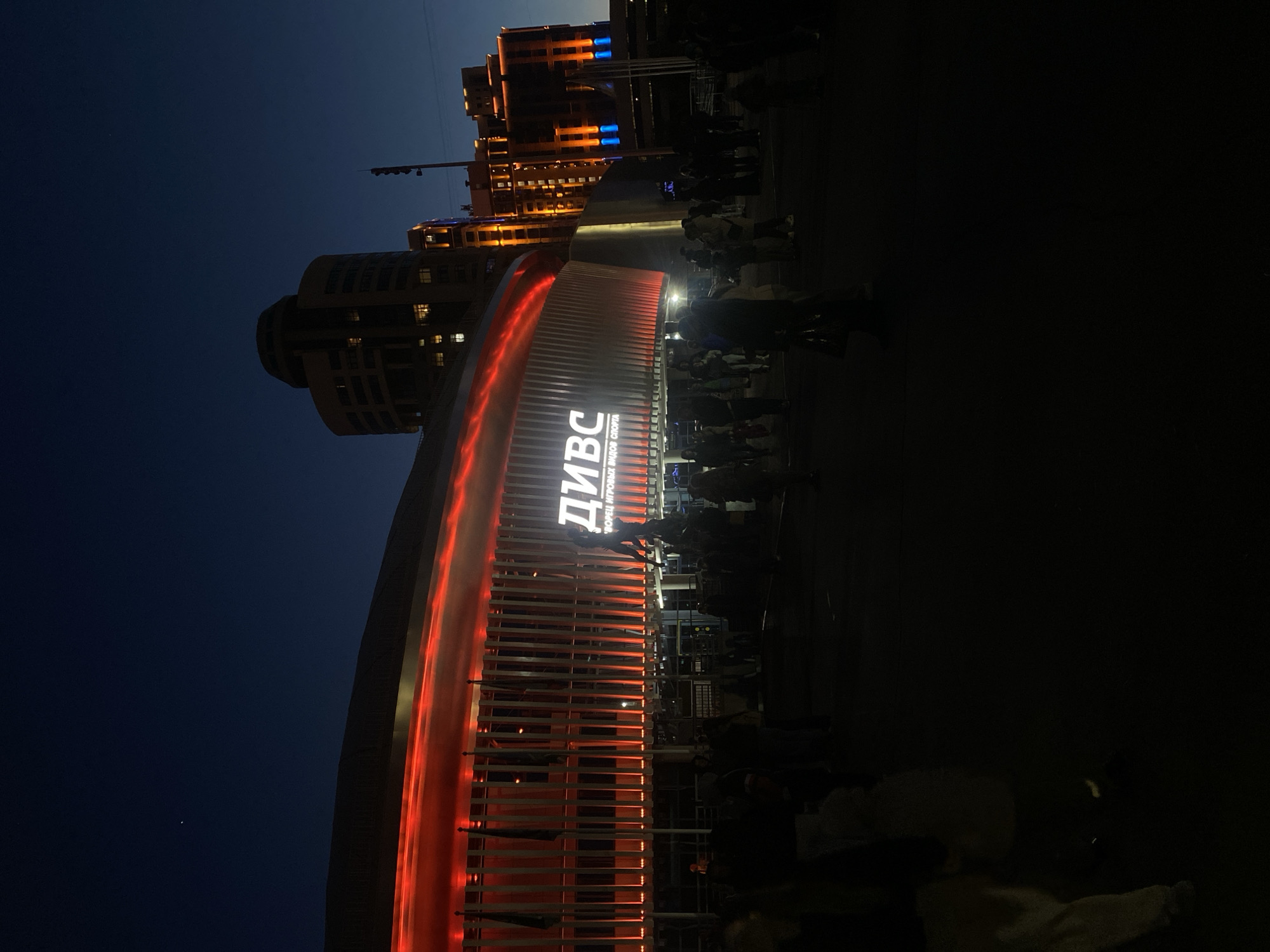 Captured at night, the image presents a view of the DIVS Uralochka building in Yekaterinburg, Russia. The building's facade is defined by a striking red arched design, and the signage on the structure is prominently displayed. Adjacent to the DIVS building is a multi-story structure illuminated with both orange and blue lights. Several individuals are present near the DIVS building, positioned around its entrance. The sky transitions from light to dark.