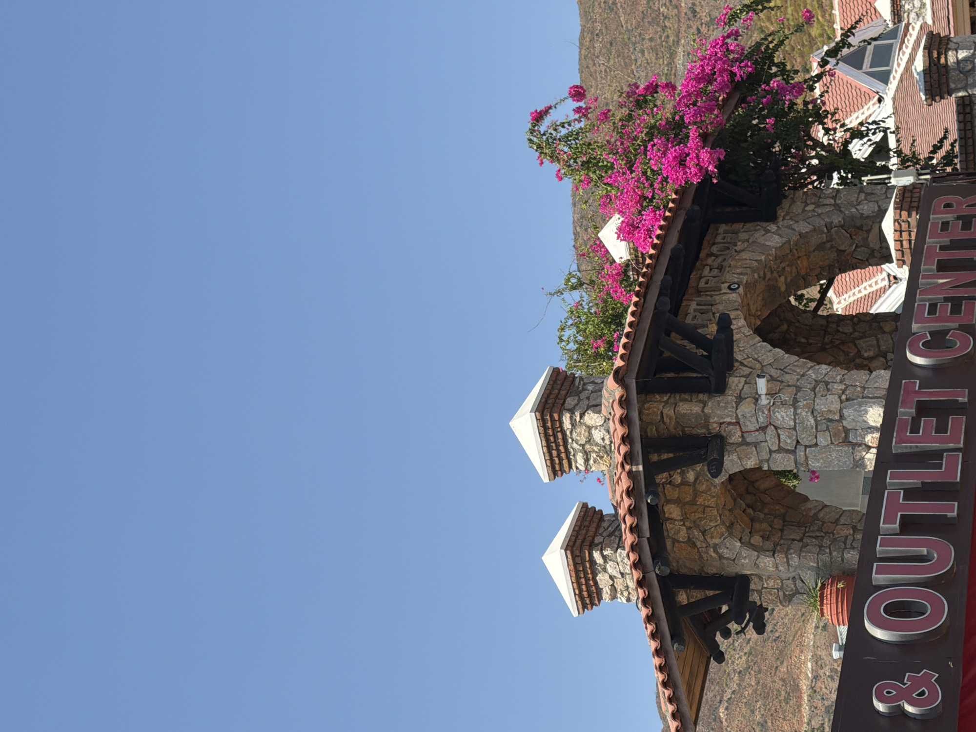 The image captures a bright, sunny day in Marmaris, Turkey, with a clear blue sky dominating the upper portion. In the foreground, a rustic stone archway with a tiled roof is adorned with vibrant pink bougainvillea flowers. Two ornate brick chimneys with white caps rise from the roofline. Below the archway, a prominent sign reads "& OUTLET CENTER" in bold, red lettering with white outlines. The architecture suggests a resort or shopping complex. The background features a dry, hilly landscape characteristic of the region. No people are visible in the frame.
