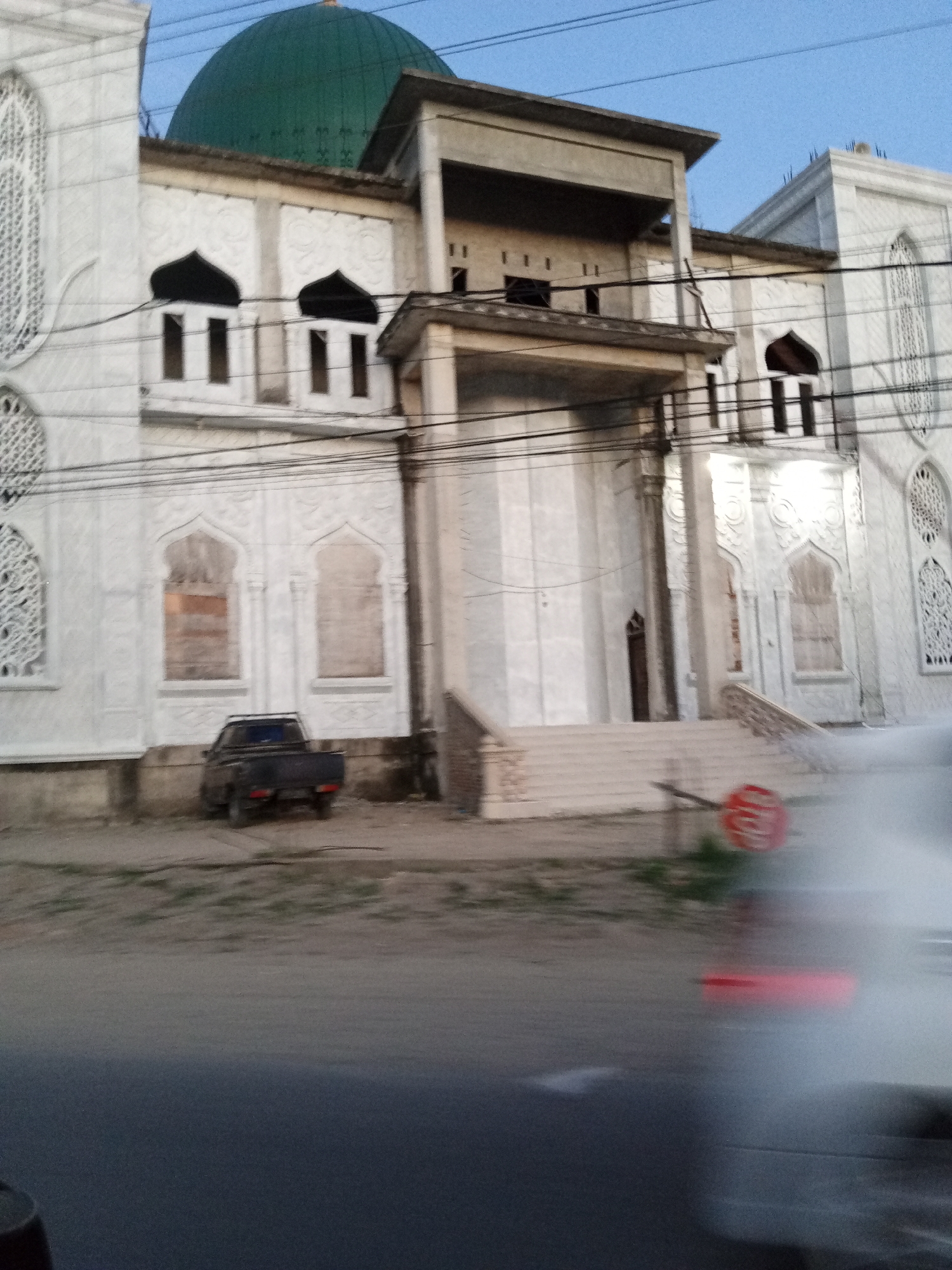 The image shows a large, white building with a green dome, identified as a mosque, located in Keudelapang, Indonesia. The building has arched windows and intricate facade detailing. In front of the building, there's a small black pickup truck parked on a dirt area. The street in the foreground is asphalt, and there's a blurry object, likely a vehicle, moving across the frame from right to left. Overhead power lines run across the front of the building.