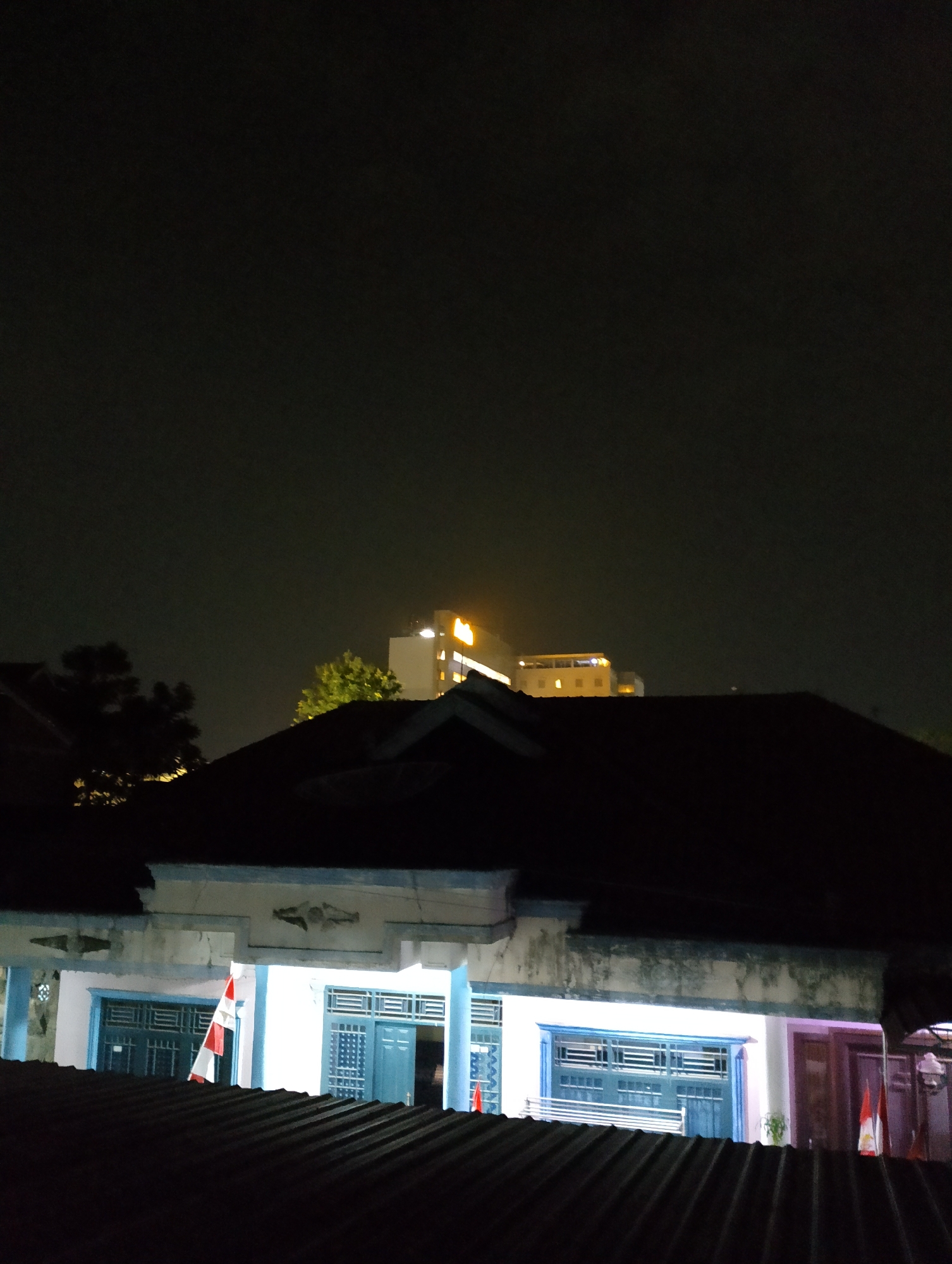 This is a night-time, medium-shot, eye-level, vertical photograph taken in Magelang, Indonesia. The foreground is dominated by the dark, corrugated metal roof of a building. Above this, a white building with blue accents is visible. This building has several blue doors with decorative metal grilles and is adorned with Indonesian flags. Further in the background, silhouetted trees are visible against a dark sky, with a brightly lit, multi-story building in the distance. The overall scene depicts a residential or commercial area at night.