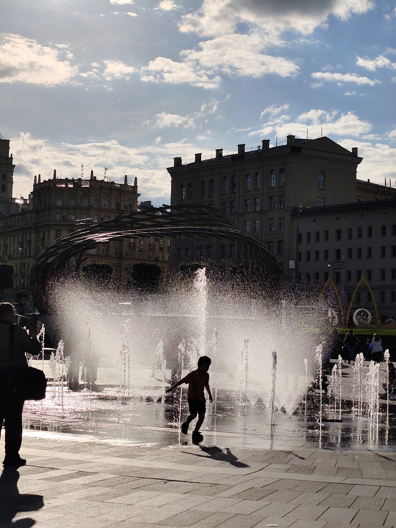 A wide-angle, outdoor shot captures a sunny day in Moscow, Russia, with a focus on a fountain area. The light is strong enough to create silhouettes, and some water droplets are suspended in the air from the fountain.
In the foreground, a young boy is silhouetted as he runs and plays in the fountain. To the left, another person, partially silhouetted, stands with a bag and appears to be taking photos. The fountain itself consists of numerous jets of water spraying upwards. The ground is paved with large, light-colored rectangular stones that show the reflections of the fountain and the sky.
In the background, a large, arched metal structure is visible, along with several buildings of various heights and architectural styles. Some people are seated on benches in the background, near another, smaller, circular structure. The sky is blue with scattered white clouds, and the sunlight is particularly strong near the top-left corner of the image.