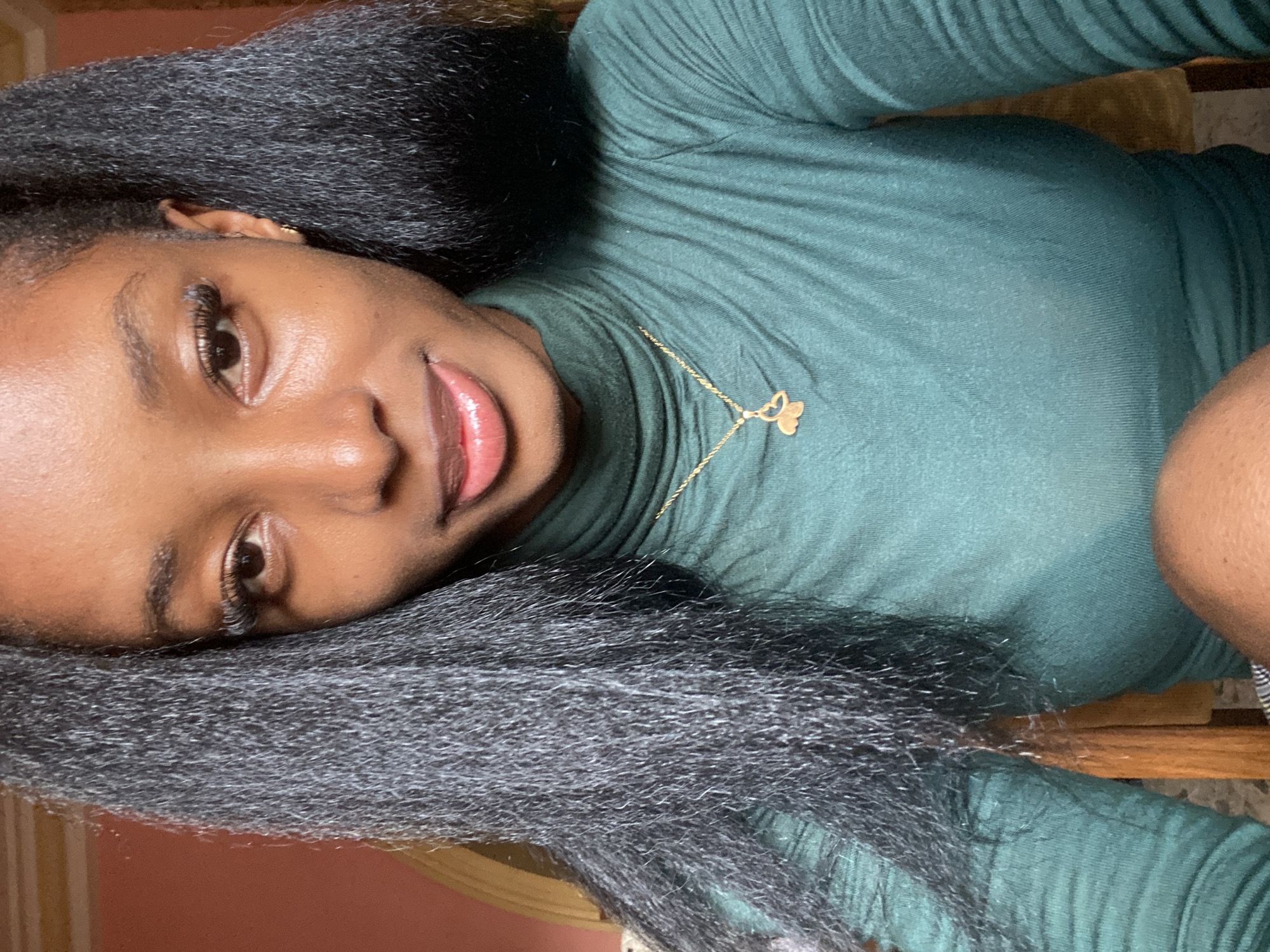 An individual, identified as female, with a dark complexion, is positioned centrally in a medium shot, gazing directly into the camera in the city of Jos, Nigeria. Her dark, coarse-textured hair is styled in two distinct sections, framing her face and extending over her shoulders. Facial features include brown eyes with pronounced eyelashes, well-defined eyebrows, and full, glossy lips slightly parted, revealing the tip of her tongue. She is wearing a dark teal, ribbed turtleneck long-sleeved top. A thin, gold-colored chain necklace with a small, butterfly-shaped pendant hangs around her neck. A small gold-colored stud earring is visible in her right earlobe. The background consists of a plain, muted reddish-brown wall and fragments of light and dark wooden furniture. The lower right portion of the image shows a segment of her bare knee.