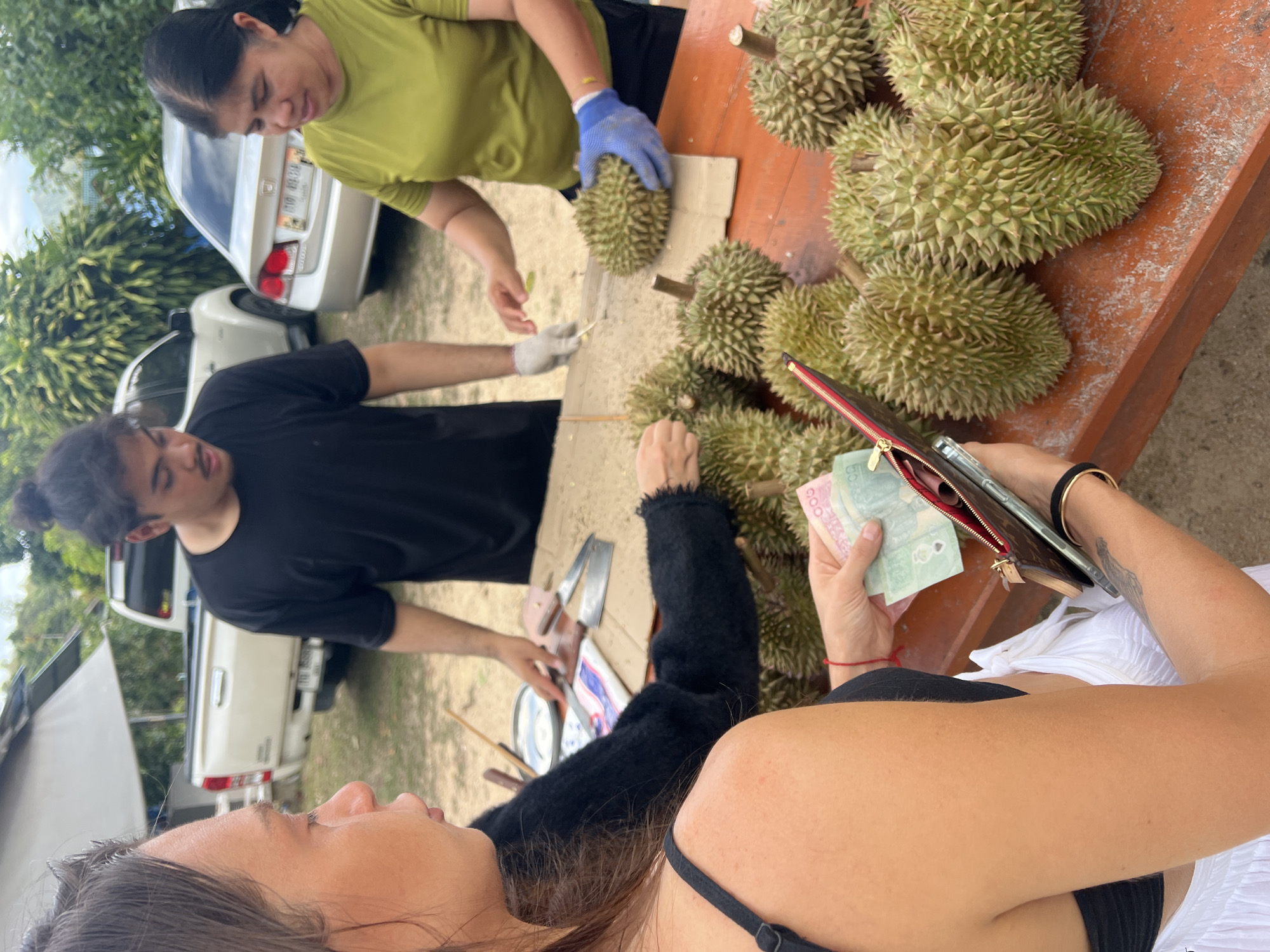 In บ้านบางรักษ์, Thailand, the scene presents two women and a man standing around a table laden with durian fruits. One woman, wearing a green shirt and blue gloves, appears to be handling the durians with a knife on a wooden surface, while the man, in a black shirt, seems to be assisting in the task with a knife. The second woman is holding Thai baht currency and a purse. A table sits underneath an awning and near a couple of automobiles.