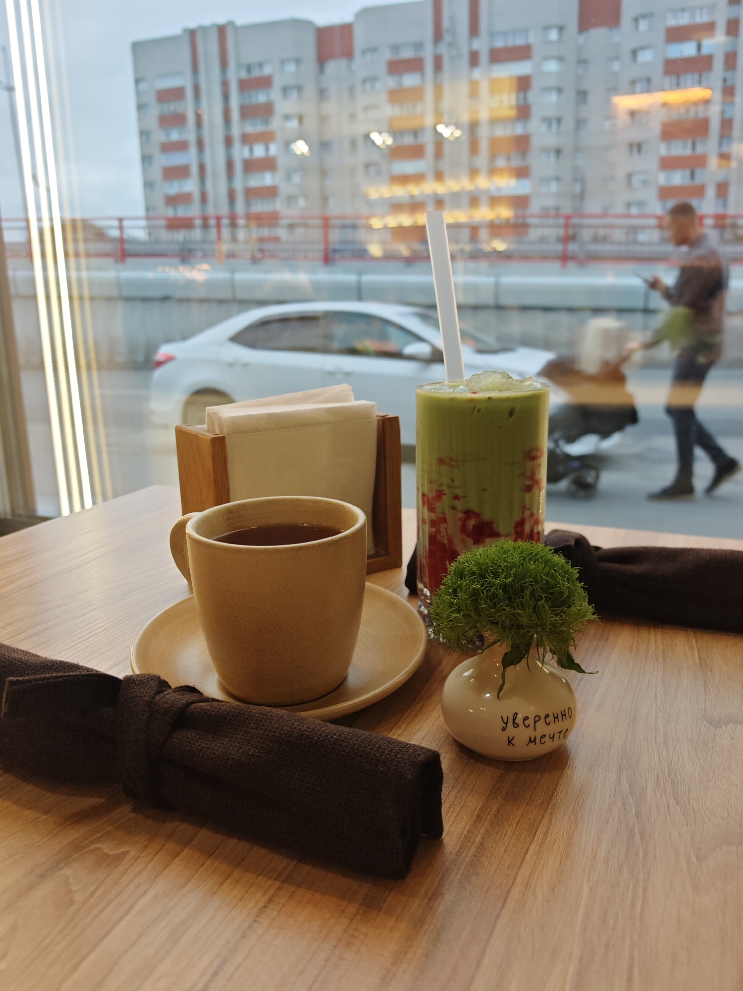 A close-up shot shows a mug of dark tea on a saucer, a green drink with red chunks and a straw, and a small vase with a green fluffy plant on a wooden table. In the background, through a window, a blurry city scene is visible, including a white car, a building with red and white facades, and a person walking. The text on the vase reads "Уверенно к мечте" which translates to "Confidently towards the dream." The scene is likely a cafe or restaurant in Novy Urengoy, Russia, given the city's distinctive architecture and the use of Russian text.