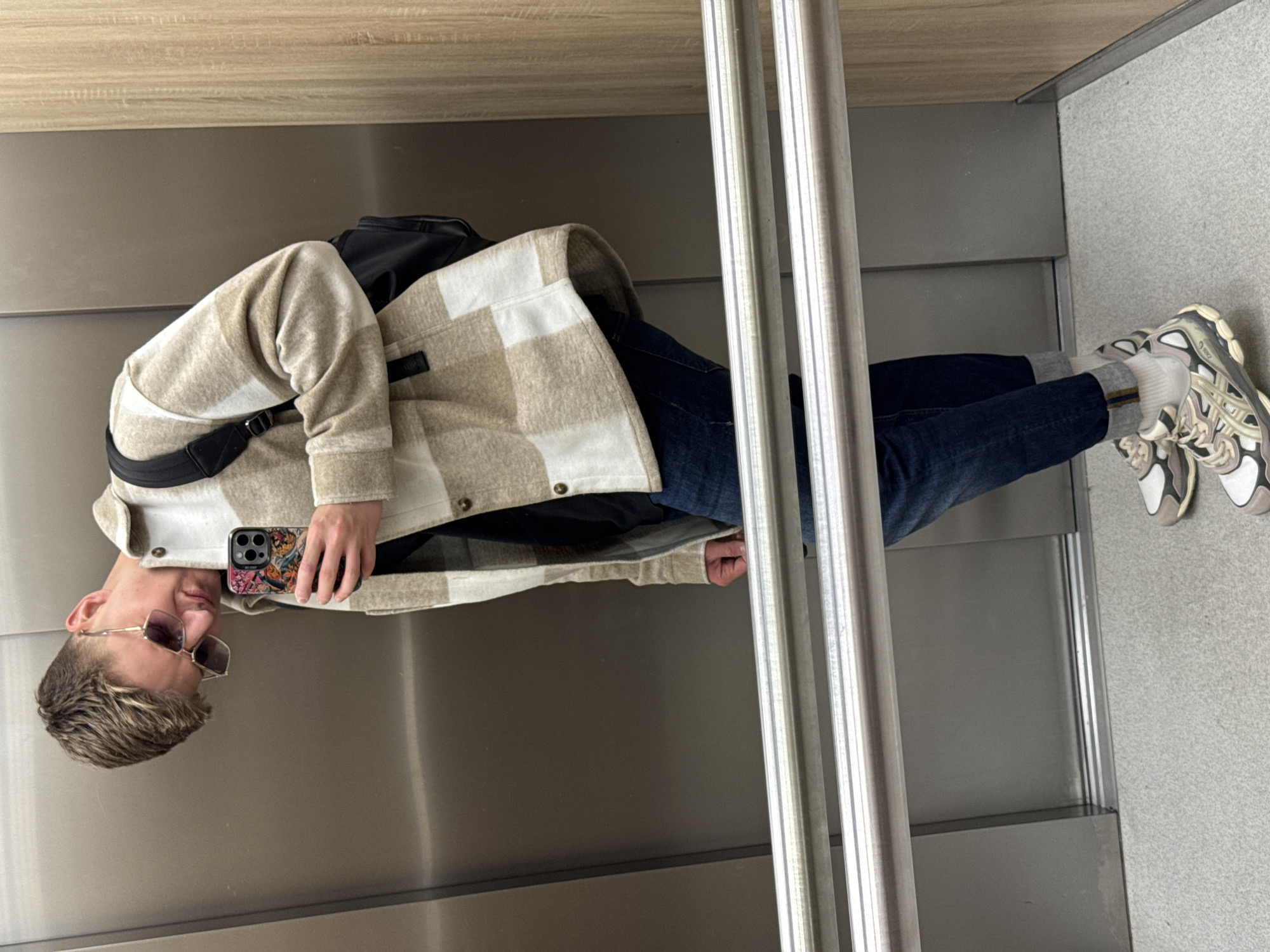 A person stands in an elevator taking a selfie. The elevator's interior is utilitarian with metal walls and flooring. The person is wearing jeans, white socks, sneakers, a black backpack strap, and a beige and white plaid jacket, and sunglasses. They are holding a phone with a colorful case. The image does not contain information about the location in Plovdiv, Bulgaria.