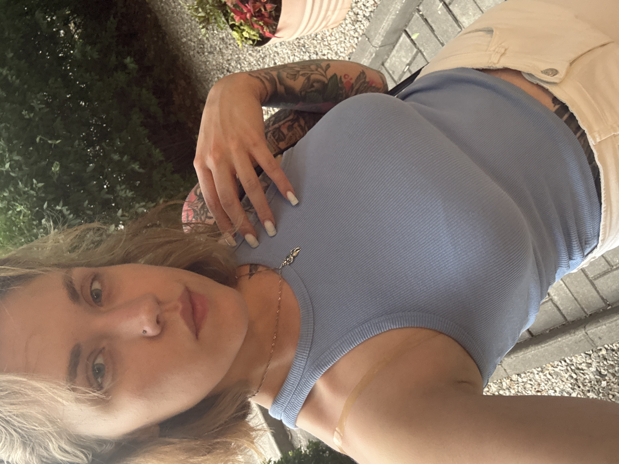 A young woman with blonde hair and tattoos on her arm poses for a selfie. She is wearing a light blue ribbed tank top and light-colored shorts. Her left hand is visible, resting on her chest. A thin gold chain with a small pendant is around her neck. The background shows greenery, gravel, and a paved walkway, suggesting an outdoor setting. The lighting indicates it is daytime. The image is taken from a high angle, looking down at the woman. There is no visible text in the image. The provided location context is Almaty, Kazakhstan.