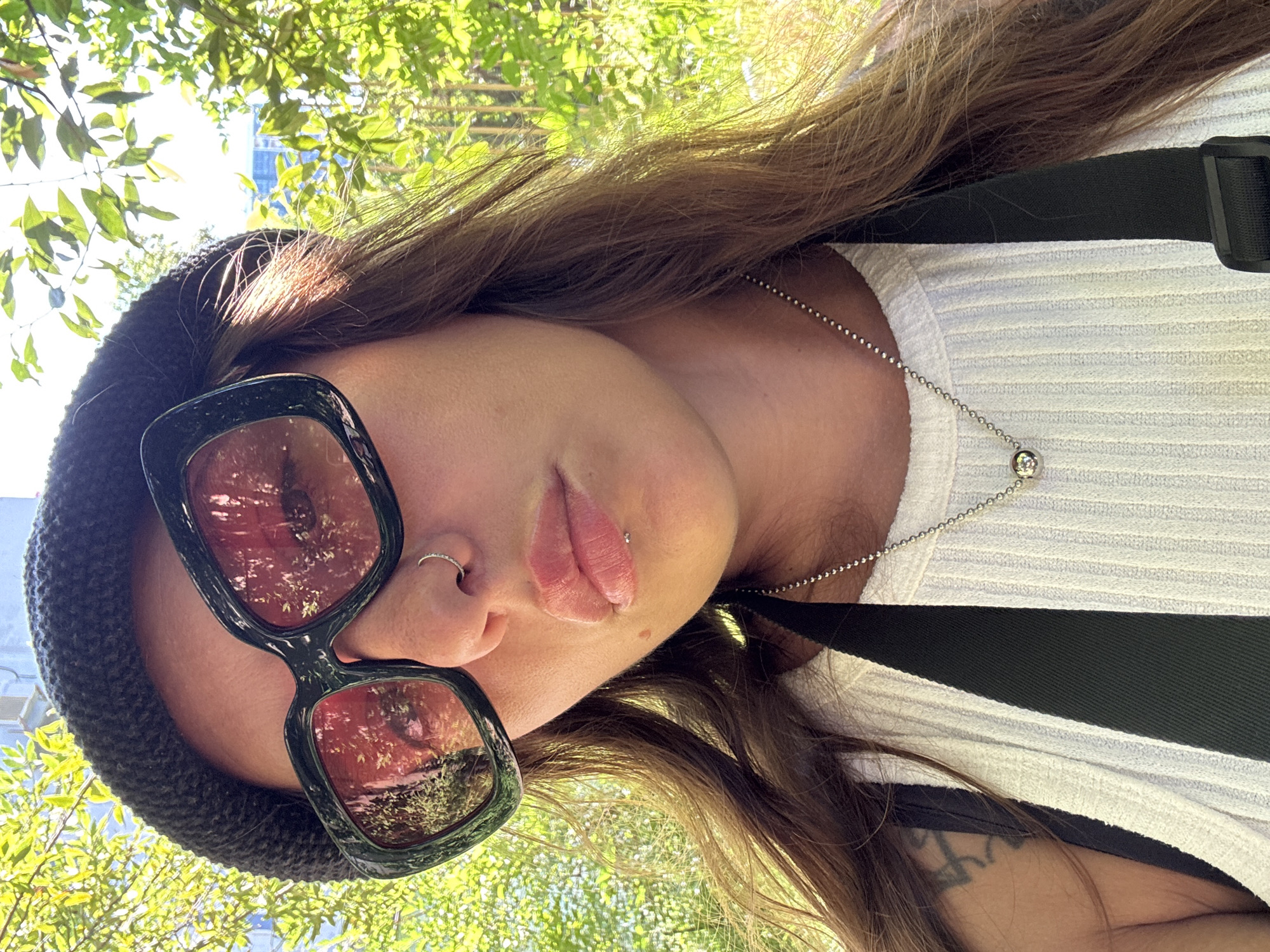 A woman is posing for a selfie outdoors, with lush green foliage and dappled sunlight in the background. She is wearing a dark knit beanie, oversized sunglasses with pink lenses, and a white ribbed tank top. A silver ball chain necklace with a round pendant rests against her chest. She has a nose ring and a lip ring. A portion of a tattoo is visible on her left shoulder. The image appears to be taken during the daytime, possibly in a park or garden setting.