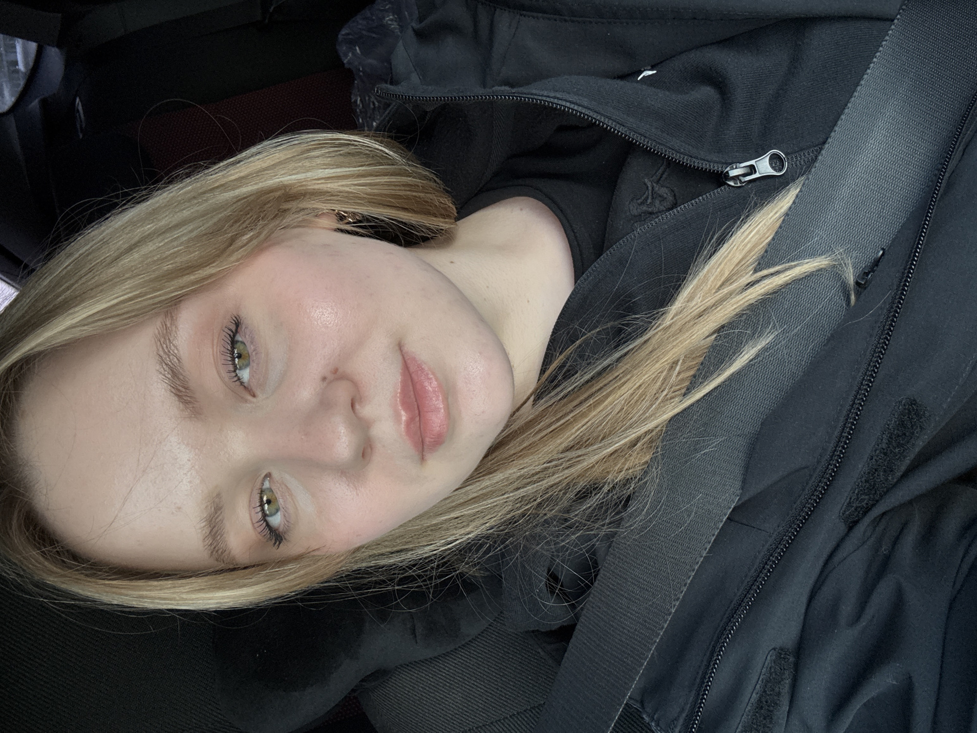 A young person, likely female, with fair skin and long, light blonde hair, is captured in a close-up, rotated portrait. The individual is positioned inside what appears to be a car or vehicle, indicated by a dark seatbelt diagonally crossing their body and a dark car interior visible in the background. The setting is in Chaikovsky, Russia.
The person is wearing a black zippered jacket over a black top. Their light green eyes are open and looking directly at the camera, conveying a neutral to calm expression. Subtle makeup, including mascara and a light pink lip color, is visible. An earbud is partially visible in their right ear. The lighting is soft and natural, though the exact time of day or weather cannot be determined from the image. No visible text is present in the frame. The image is composed at an angle, giving it a dynamic feel.