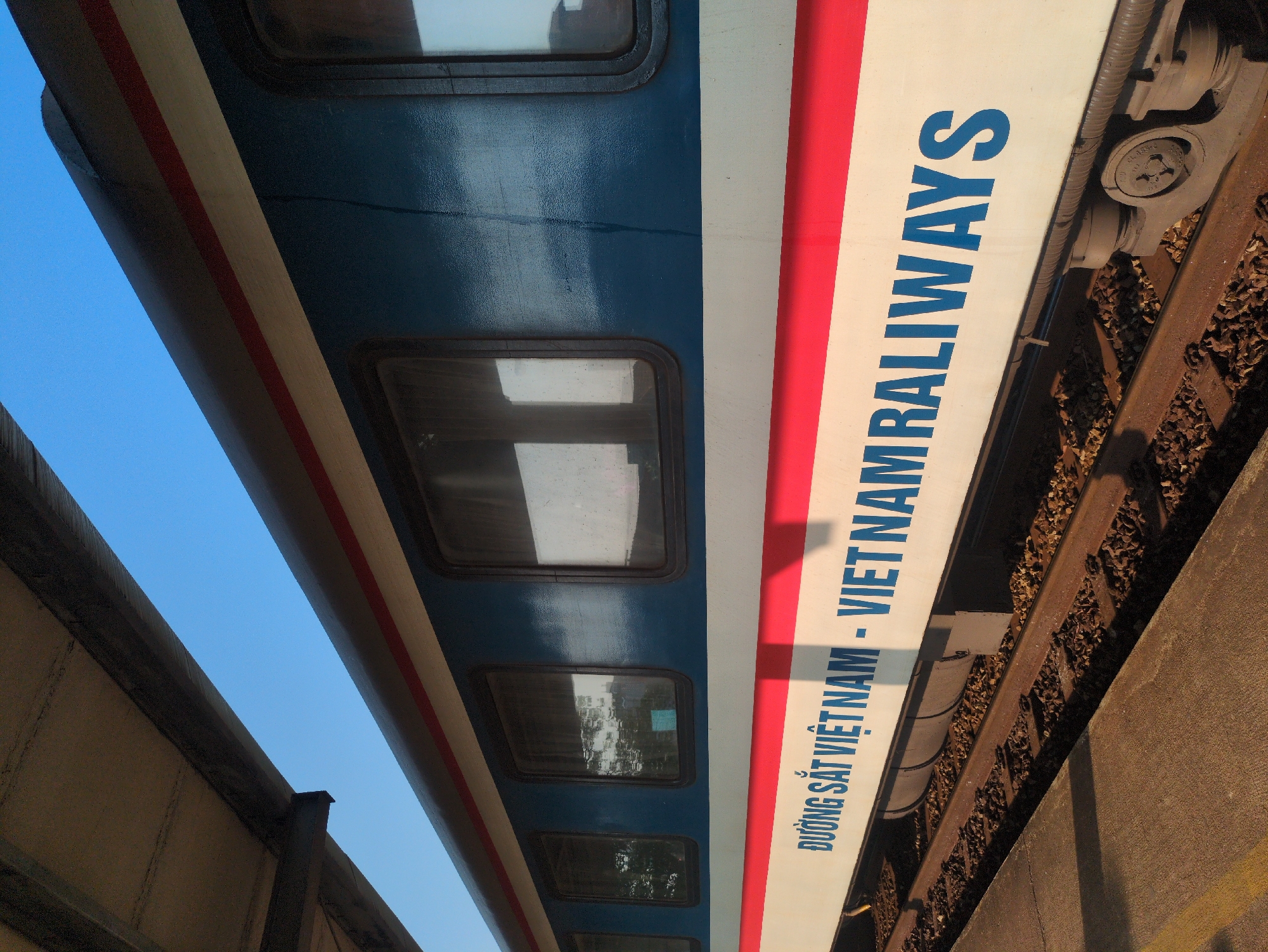 The image captures a segment of a Vietnam Railways passenger train in Tuy Hòa, Vietnam. The train carriage is predominantly blue with a horizontal white stripe bordered by red. "VIETNAM RAILWAYS" is printed in large, blue, sans-serif font on the white stripe, with smaller Vietnamese script above it. The train is positioned on railway tracks composed of brown wooden ties and rust-colored metal rails. A concrete platform is visible alongside the tracks. Windows with black frames are present on the blue section of the train car. The background reveals a clear blue sky. The shot appears to be taken from a low angle looking upward, and the scene is well-lit.