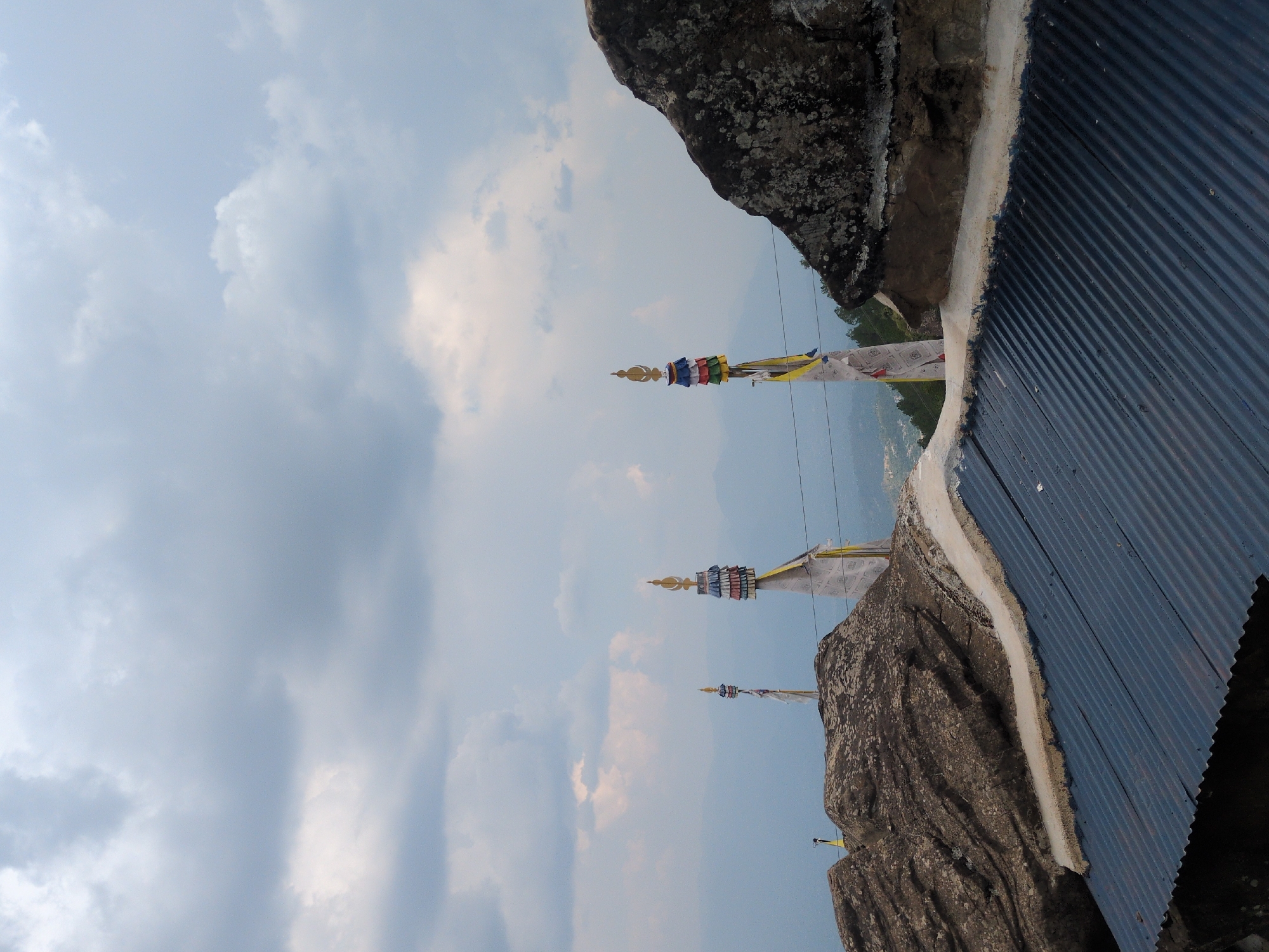 This image depicts a mountainous landscape with a cloudy sky. In the foreground, a corrugated metal roof is visible, suggesting a building or structure. Beyond the roof, large rock formations dominate the scene, some appearing weathered. Several Buddhist prayer flags are mounted on poles, with colorful fabric sections and intricate finials. The flags are positioned at various heights and distances, adding a spiritual element to the natural setting. In the background, misty mountains recede into the distance, with hints of a valley or settlement at their base. The overall atmosphere is serene and contemplative, typical of high-altitude religious sites. The location is specified as the city of Doh, India.