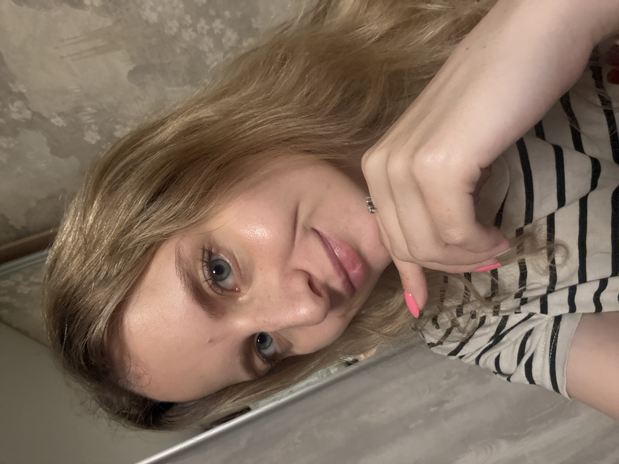 A young Caucasian woman with blonde hair and blue eyes is captured in a close-up, selfie-style photograph, seemingly taken indoors. She is lying down, with her head tilted and resting on her hand, which is adorned with bright pink nail polish. Her expression is relaxed and she has a slight smile. She is wearing a striped shirt. The background is blurred, but appears to be a wall with a textured, possibly wallpapered, pattern. The lighting suggests it could be evening or an indoor setting with artificial light. The overall mood is casual and intimate.