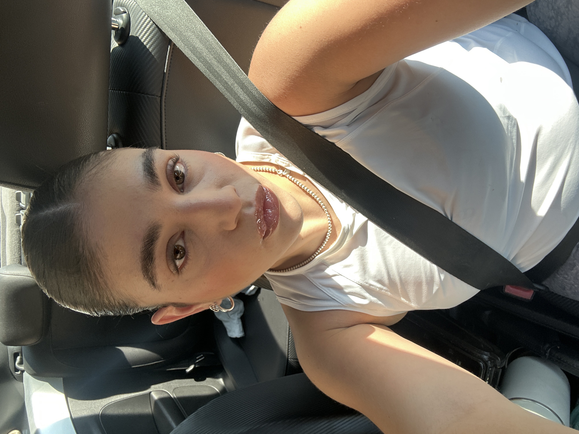A woman is in the passenger seat of a car. She is wearing a white top and a silver necklace, and has on a seatbelt. Her hair is slicked back. The interior of the car is black leather. There is a water bottle on the floor of the car. The location is unknown and cannot be determined from this photo.