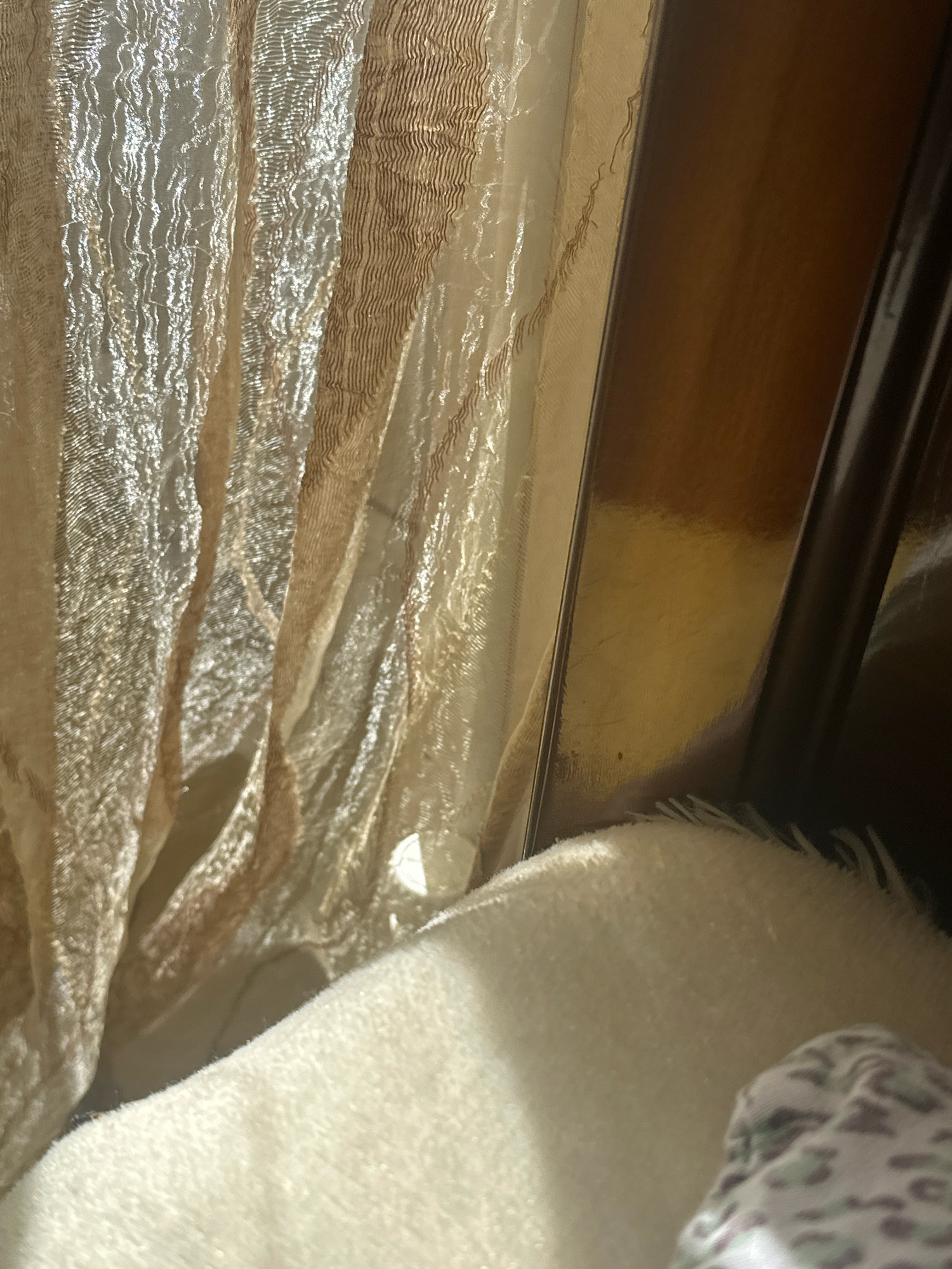 The image shows a close-up of draped fabric, likely curtains, with a textured, shimmering surface in shades of beige and gold. The fabric is caught by sunlight, creating highlights and shadows that emphasize its crinkled appearance. To the right of the fabric, a polished wooden surface is visible, reflecting light. In the foreground, a soft, fluffy white blanket is partially visible, with a hint of a patterned fabric, possibly a blanket or clothing, peeking out from the lower right corner. The overall impression is one of indoor comfort, with natural light playing on textiles. There are no discernible features that would indicate a specific location within the city of Gyumri, Armenia.