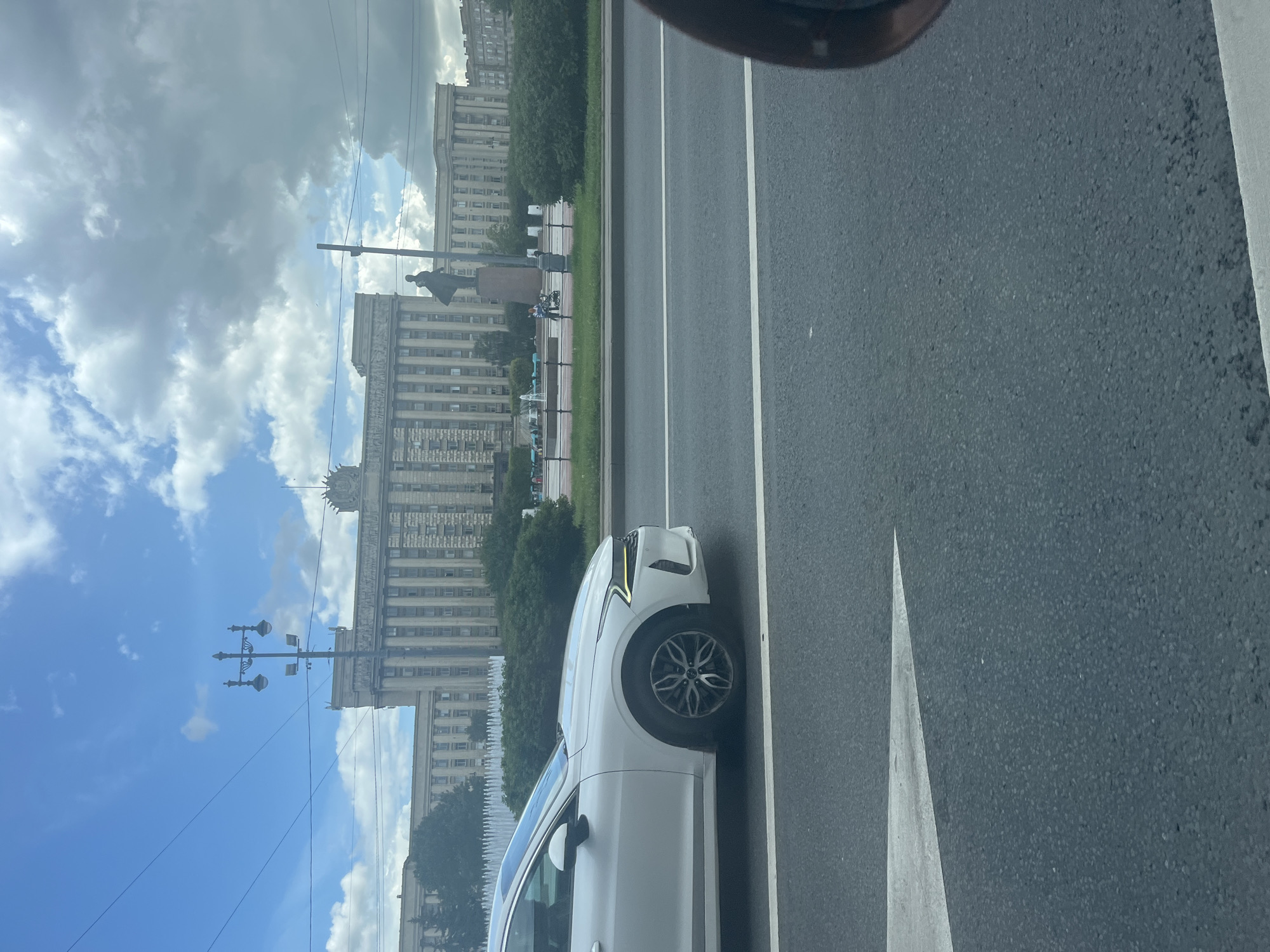 A white Kia K5 is captured in motion from the side, traveling on a multi-lane road in Saint Petersburg, Russia. In the background, a monumental neoclassical building, likely an administrative or government structure, with a long facade of columns and a symmetrical design, stands under a cloudy sky. A statue of Vladimir Lenin is prominently situated in front of the building. The foreground is dominated by the asphalt road with white lane markings, including a large white arrow pointing to the left. A hint of another vehicle's side mirror is visible in the lower right corner of the frame. The scene suggests a typical urban environment with significant historical architecture.