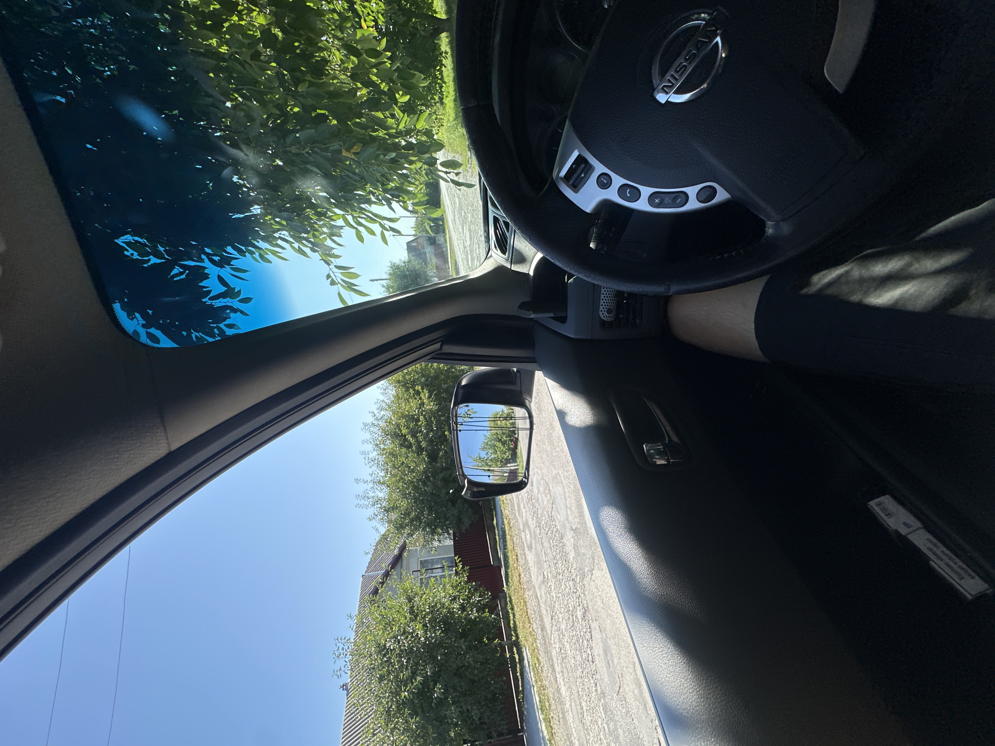 The image captures the interior of a Nissan car from the driver's perspective, showing the steering wheel, dashboard, side mirror, and a view of the exterior through the side window. Outside, a residential street with houses and trees is visible under a clear blue sky. The side mirror reflects the street and houses. A person's leg, wearing shorts, is visible in the foreground. The location appears to be Shatava, Ukraine, given the presence of what looks like typical Ukrainian village architecture. The scene is set on a sunny day.